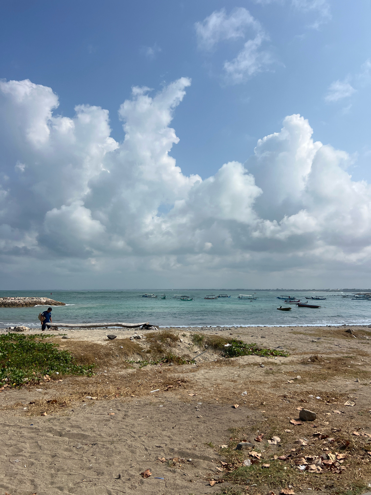 The image depicts a coastal scene in Kuta, Indonesia. In the foreground, a sandy beach is sparsely covered with dry grass, leaves, and some scattered debris, suggesting a natural, unkempt state. A large piece of driftwood lies horizontally on the sand, separating the foreground from the middle ground. In the middle ground, a person dressed in blue walks along the shoreline, carrying a bag over their shoulder. The person appears to be moving towards the left of the frame. Further along the coast, a rocky breakwater extends into the sea. The sea itself is a vibrant turquoise, dotted with numerous small, traditional Balinese boats, some of which are anchored. The water is relatively calm with gentle waves reaching the shore. The background is dominated by a bright blue sky filled with large, puffy white clouds. The overall impression is a serene day at the beach with signs of local maritime activity.