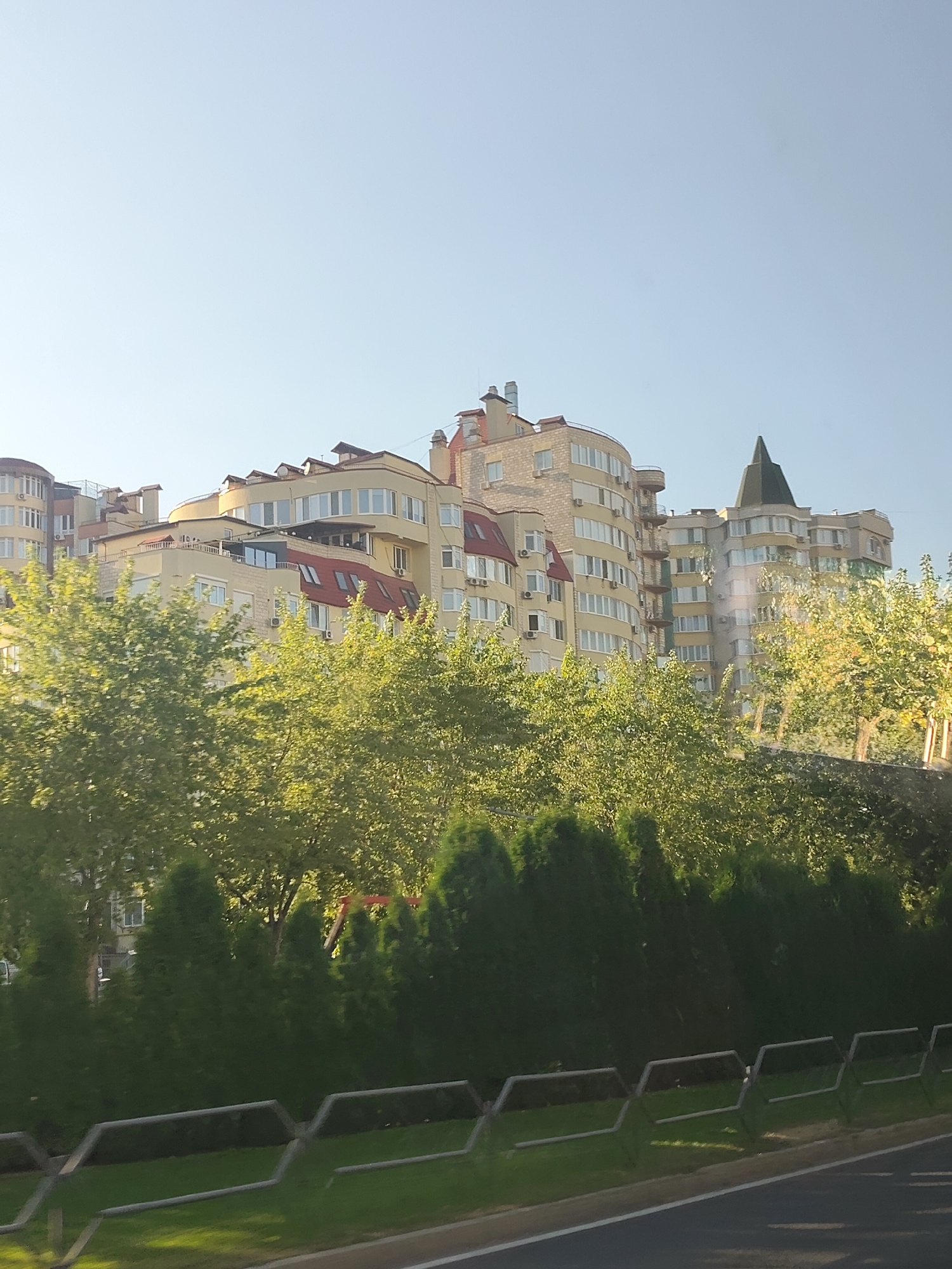 The image shows a modern apartment building complex on a clear day. The building has a curved facade, light yellow walls, and numerous windows with white frames. It features several rooflines, some with red tiles and dormer windows, and a prominent dark green, pointed turret on one section. Trees and manicured bushes line the front of the building, with a metal fence separating them from a road or pathway. The sky is a pale blue. The architecture suggests a contemporary residential area in a city, possibly Chișinău, Moldova, given the mention of that location. There are no people visible in the image.