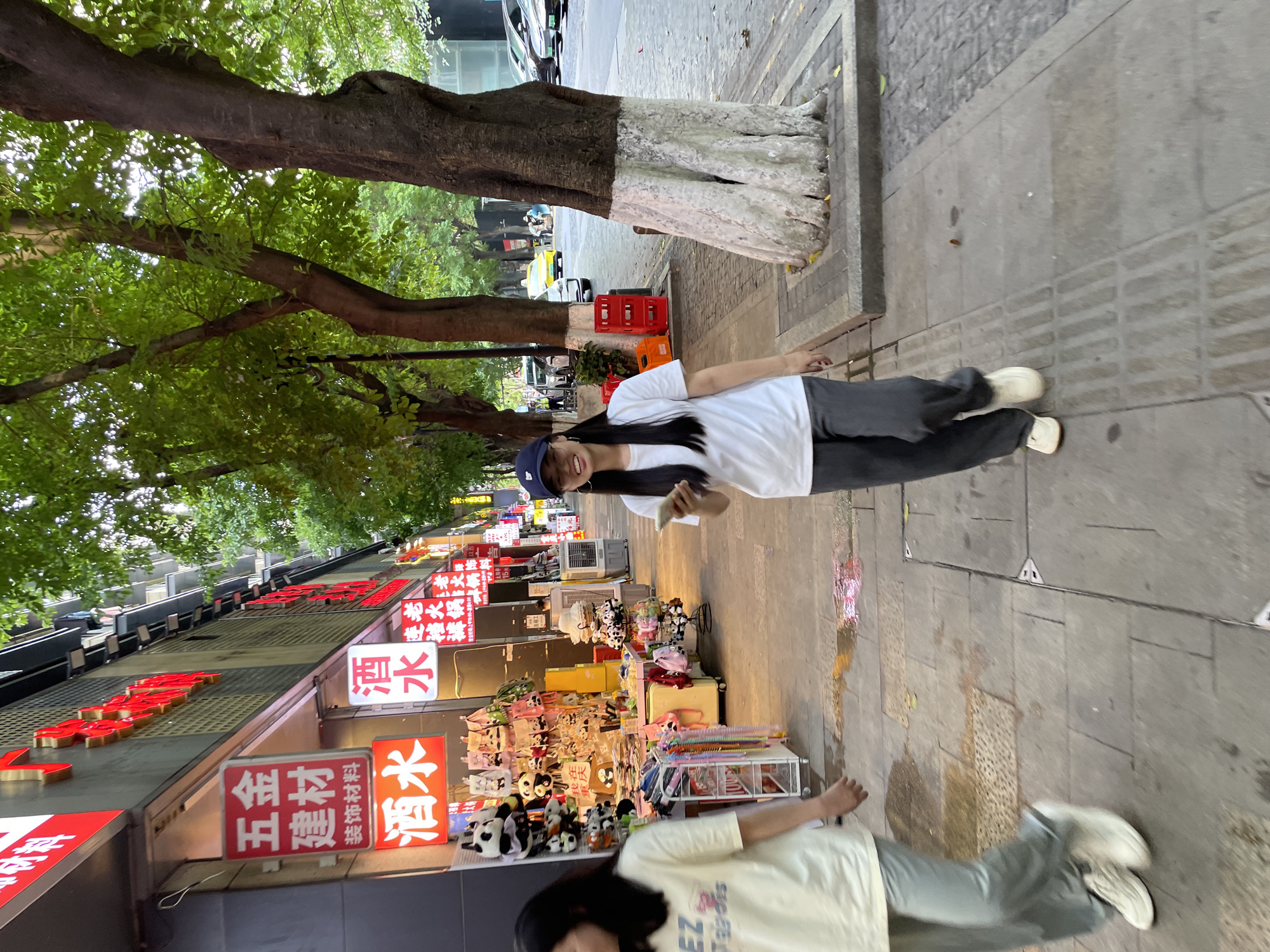 A young woman wearing a white t-shirt, grey pants, and a dark baseball cap is walking on a sidewalk while looking at her phone. Another young woman with long dark hair, wearing a light yellow t-shirt and light grey pants, is walking ahead of her on the same sidewalk. The sidewalk is paved with stone slabs and has tactile paving for the visually impaired. Several trees with white-painted bases line the sidewalk. In the background, there are shops with illuminated signs, some of which display Chinese characters. A yellow taxi and other vehicles are visible on the street. The scene appears to be a street in Yuzhong District, China, likely during the late afternoon or early evening.