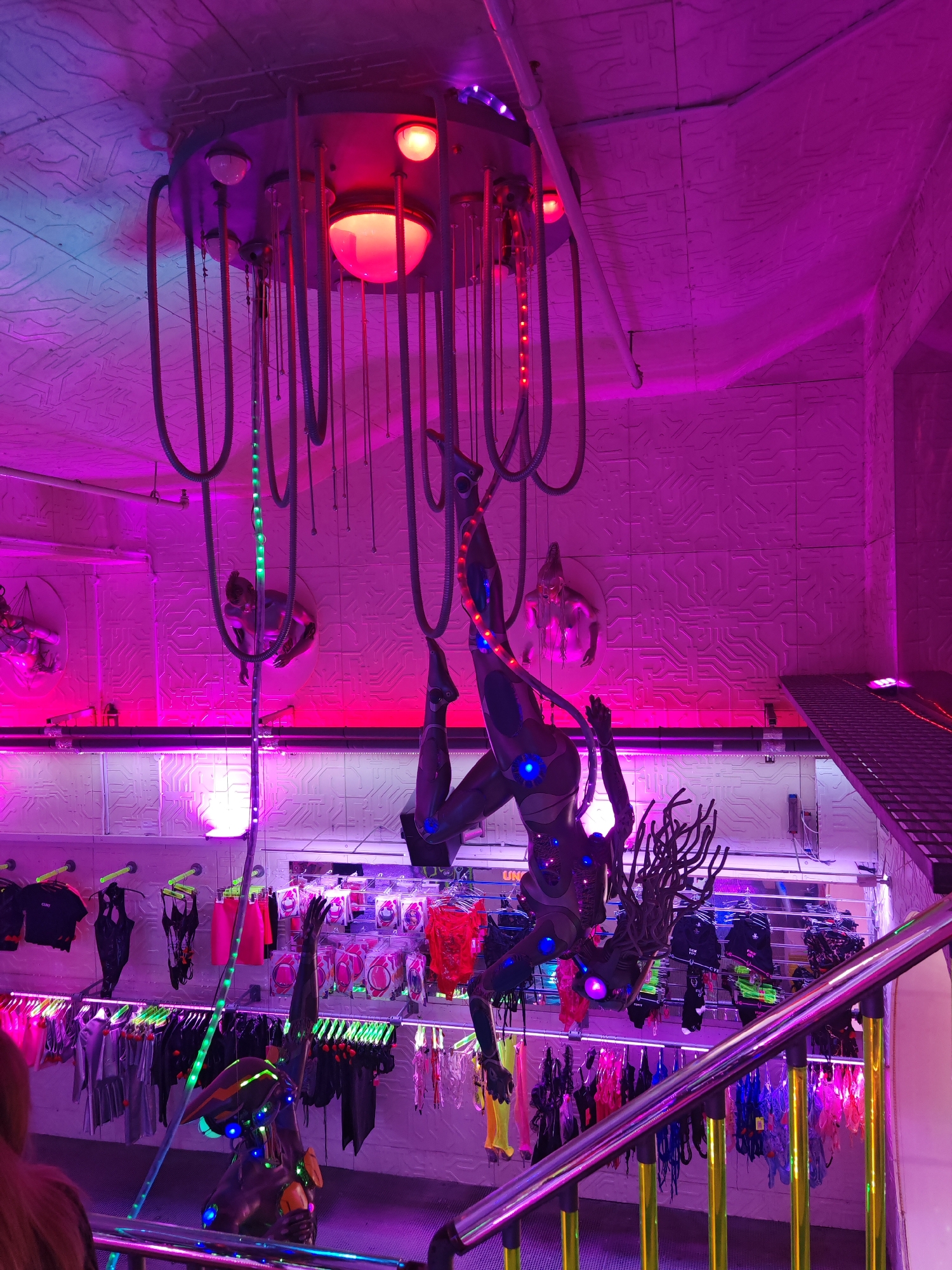 Captured in London, United Kingdom, the scene depicts an elaborately decorated interior space illuminated with vibrant, predominantly purple and pink neon lighting. A steampunk-esque installation dominates the ceiling, featuring metal tubing, illuminated globes, and a central, multi-bulb light fixture. Beneath this structure, two mannequin-like figures are suspended, appearing to be engaged in an acrobatic display. The walls are covered in patterned tiles, and a series of mannequins wearing various items of clothing are displayed on racks. A staircase with a metal railing is visible in the foreground, contributing to the complex and eclectic aesthetic of the scene.