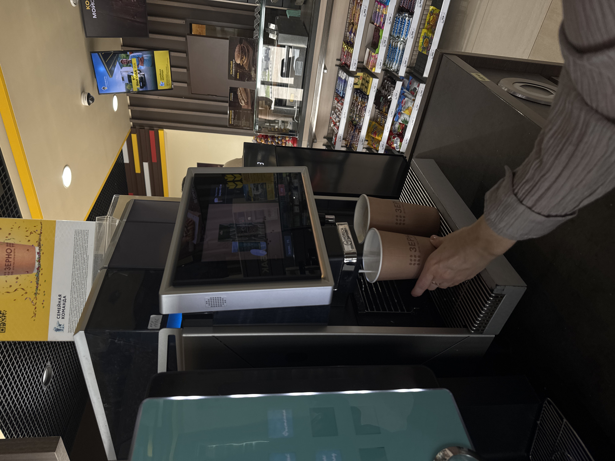 The image depicts an interior scene, likely at a gas station convenience store, as suggested by the stocked shelves of snacks and beverages visible in the background. In the foreground, a person's hand is shown reaching for or holding a paper cup under a beverage dispensing machine. Two paper cups are visible on the machine's drip tray, with one appearing to be actively filling with a beverage. The machine itself is metallic and modern, featuring a touchscreen interface. On the wall behind the machine, there are advertisements and signage. One sign, in Russian, appears to promote a "Family Team" initiative and a coffee brand, "ZERNO." The overall environment is clean and well-lit, suggesting a commercial setting. The specific location in Kochubeyevskoye, Russia is inferred from contextual information not directly present in the image itself.