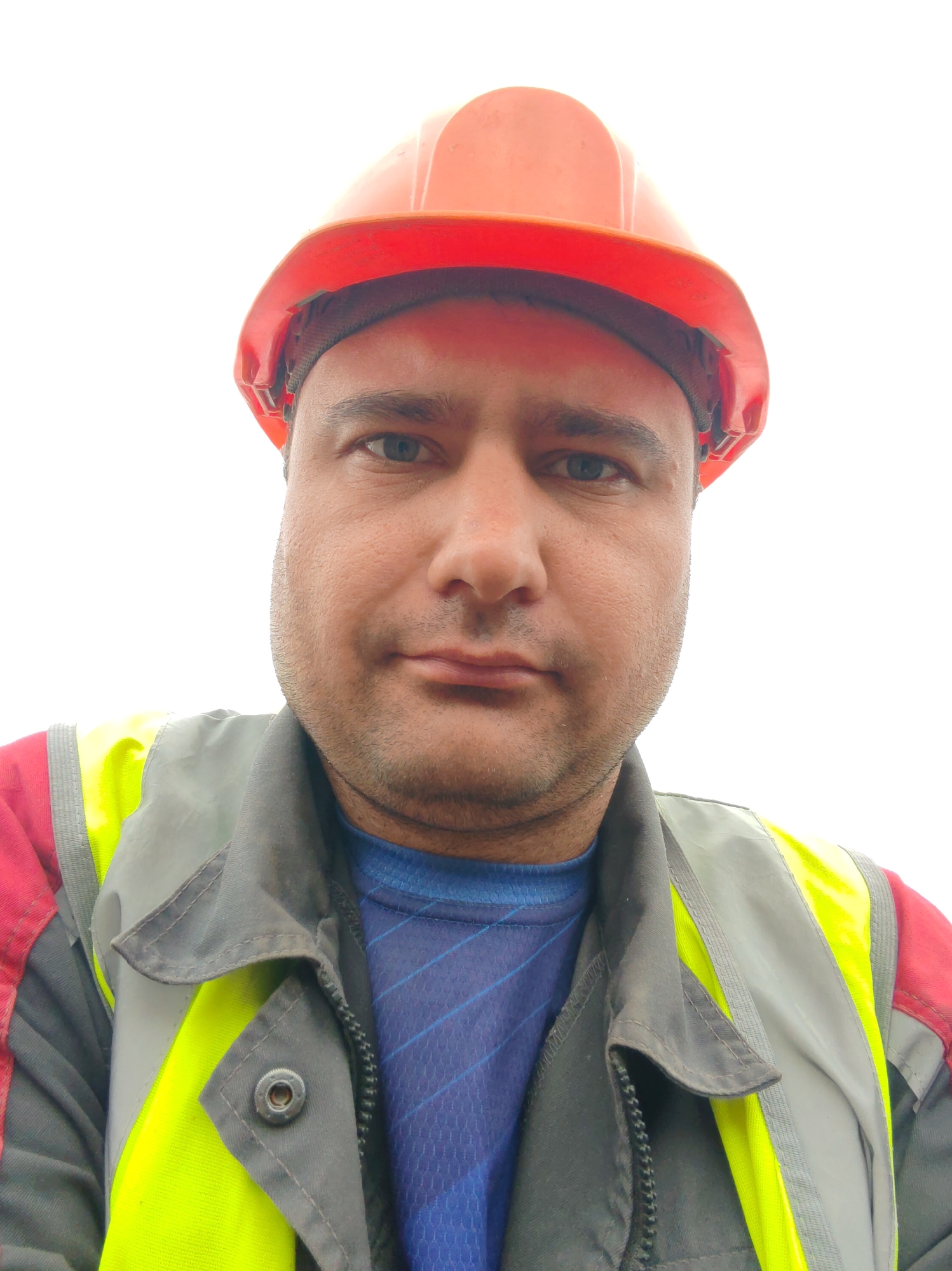 A man wearing an orange hard hat and a high-visibility vest over a blue shirt is shown in a close-up, front-facing portrait. The man has brown eyes, short dark hair, and stubble. The background is a bright white, indicating an outdoor setting, possibly a construction site or an area with strong overhead lighting. The man's gaze is directed towards the camera, and his expression is neutral. The image appears to be a selfie, taken from a low angle. The details of the location within the city are unknown.