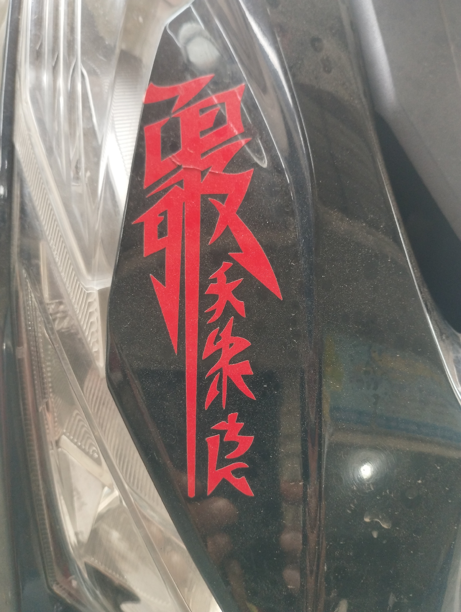 The image shows a close-up view of a sticker on a motorcycle headlight. The sticker is red and depicts Chinese characters. The characters are arranged vertically, with the top character being larger than the others. The background is a dark color, likely black. The headlight is partially visible on the left side of the image. The image does not contain any information about the location in the city of Lhokseumawe, Indonesia, or any other details related to people, subjects, objects, actions, interactions, or the environment.