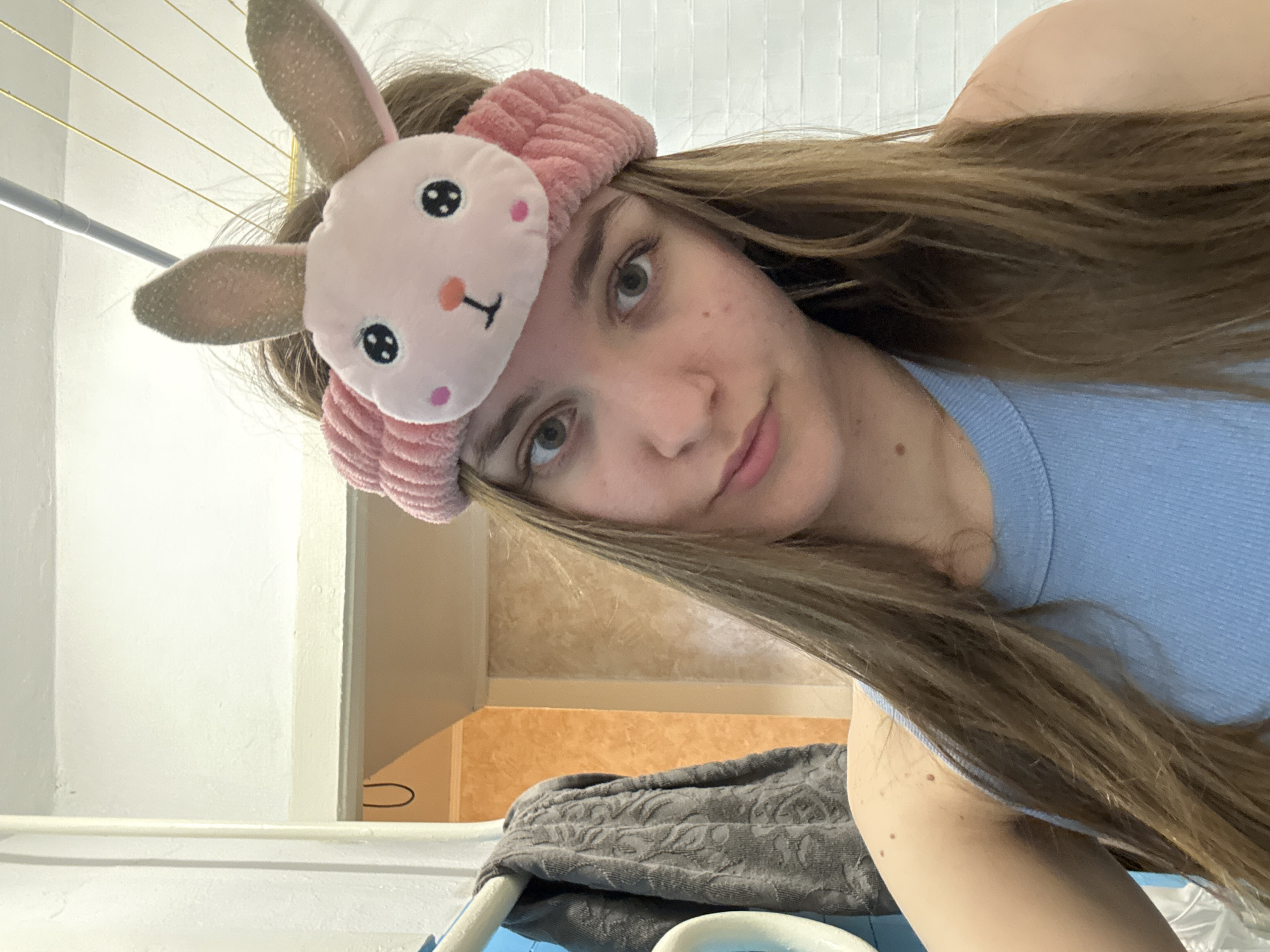 A young woman is taking a selfie indoors, wearing a light blue tank top and a pink bunny ear headband. She has long, straight, light brown hair. Behind her is a white wall, a gray patterned towel, and what appears to be a laundry basket. There are also some clotheslines visible in the background. The location in the city of Perm, Russia cannot be determined from the visual information provided.