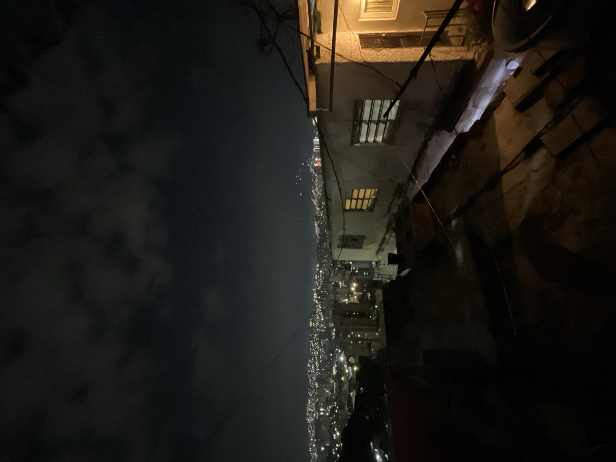 The image shows a nighttime view of Caracas, Venezuela from a high vantage point. On the right side of the image are the exterior of buildings with windows and weathered walls. On the left side of the image is a dark sky with clouds. The city lights of Caracas stretch into the distance, appearing as a dense cluster of bright points.