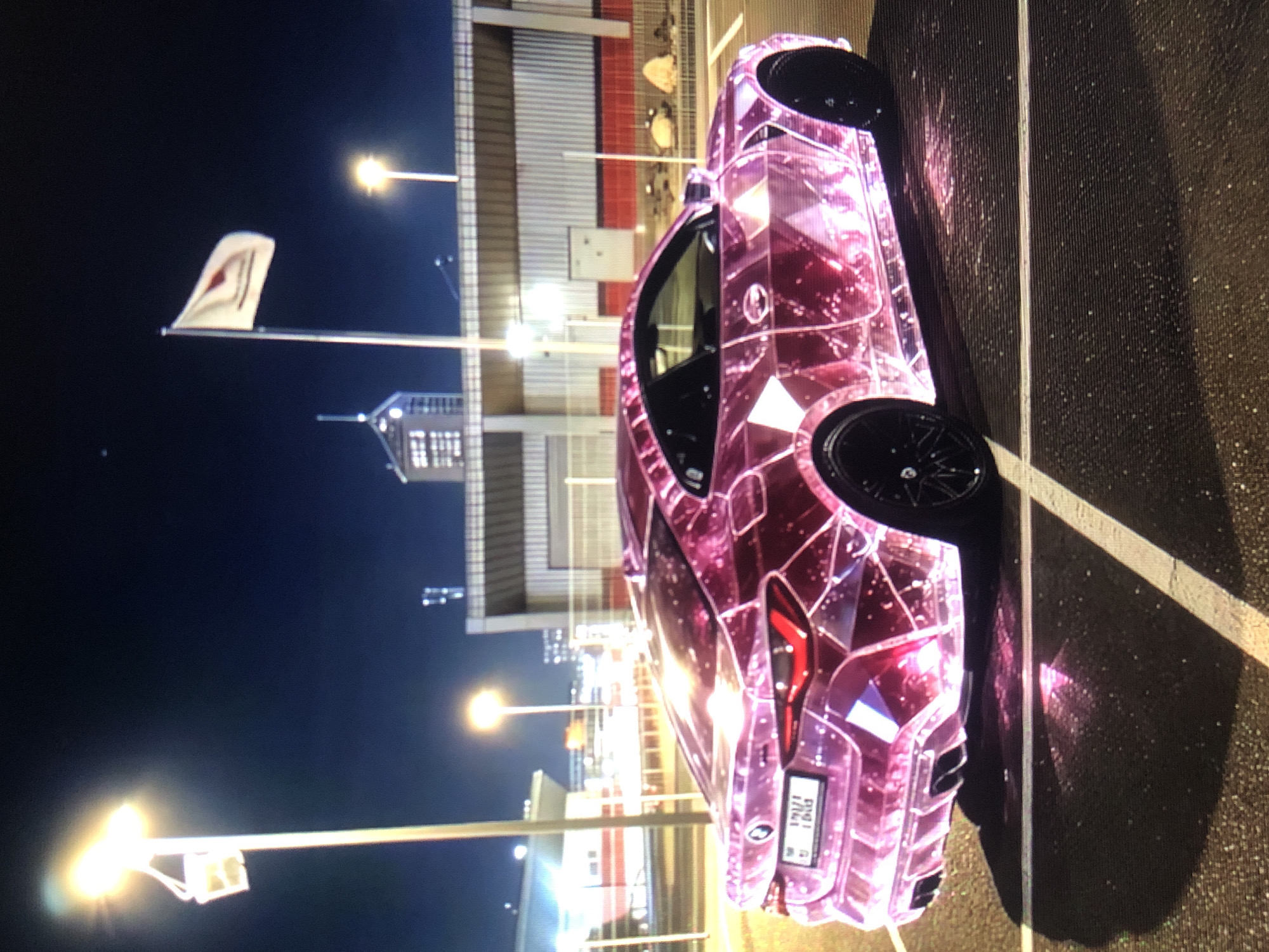 At night in Lhokseumawe, Indonesia, a pink, intricately-patterned car is parked in a marked parking space. A flag is visible in the background. Streetlights illuminate the scene.