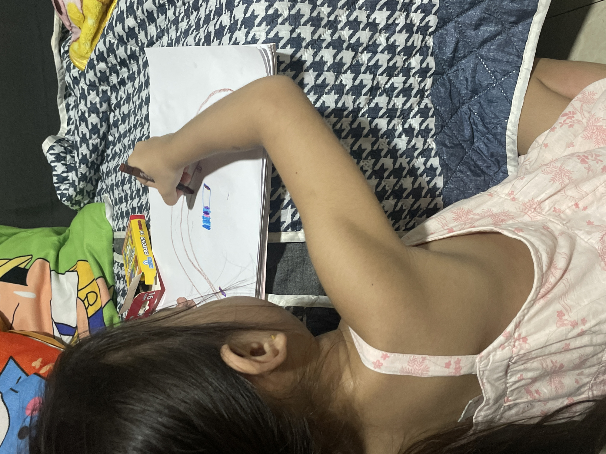 The image shows a young girl from the back, wearing a pink floral dress, drawing on a piece of paper with a brown crayon. She is sitting on a bed with a dark blue and white houndstooth patterned comforter. To her left, a box of crayons is visible. The background includes cartoon-themed pillows. The scene appears to be indoors, likely in a residential setting. The child's focus is on her drawing, and her arm is bent as she colors. The lighting suggests an indoor setting, possibly with artificial light. No specific location in Davao City, Philippines can be determined from the visual information provided.