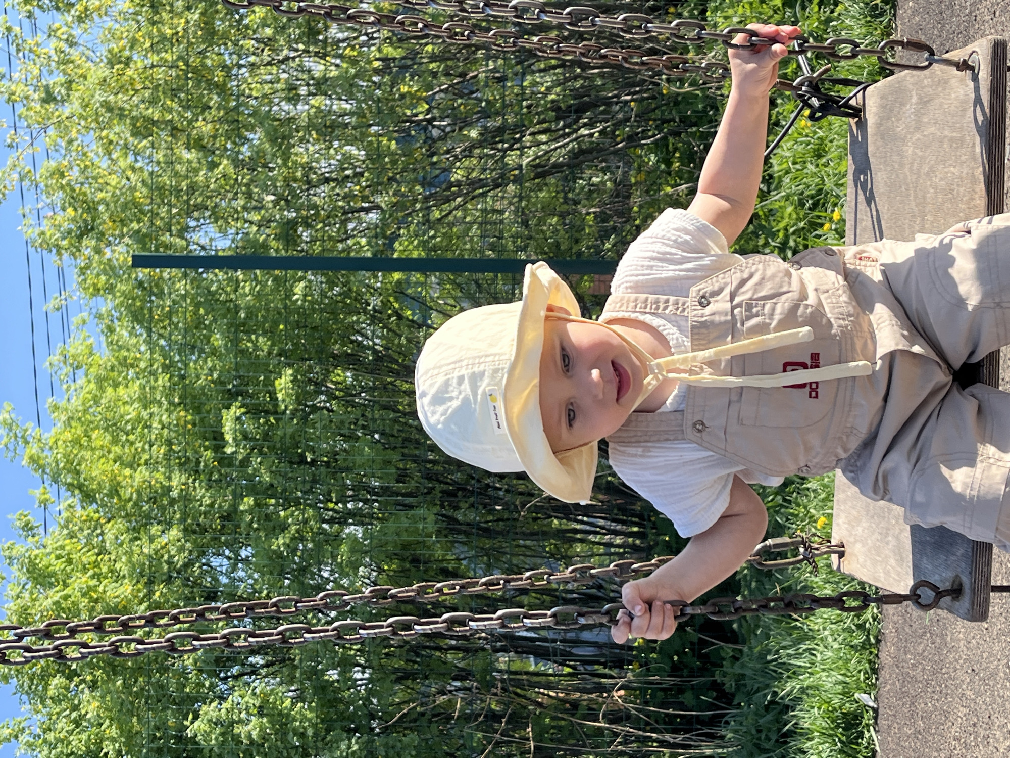 A small child wearing a light-colored sun hat and beige overalls is sitting on a swing, holding onto the chains. The swing is in an outdoor area with trees and grass visible in the background. The location is Fokino, Russia.