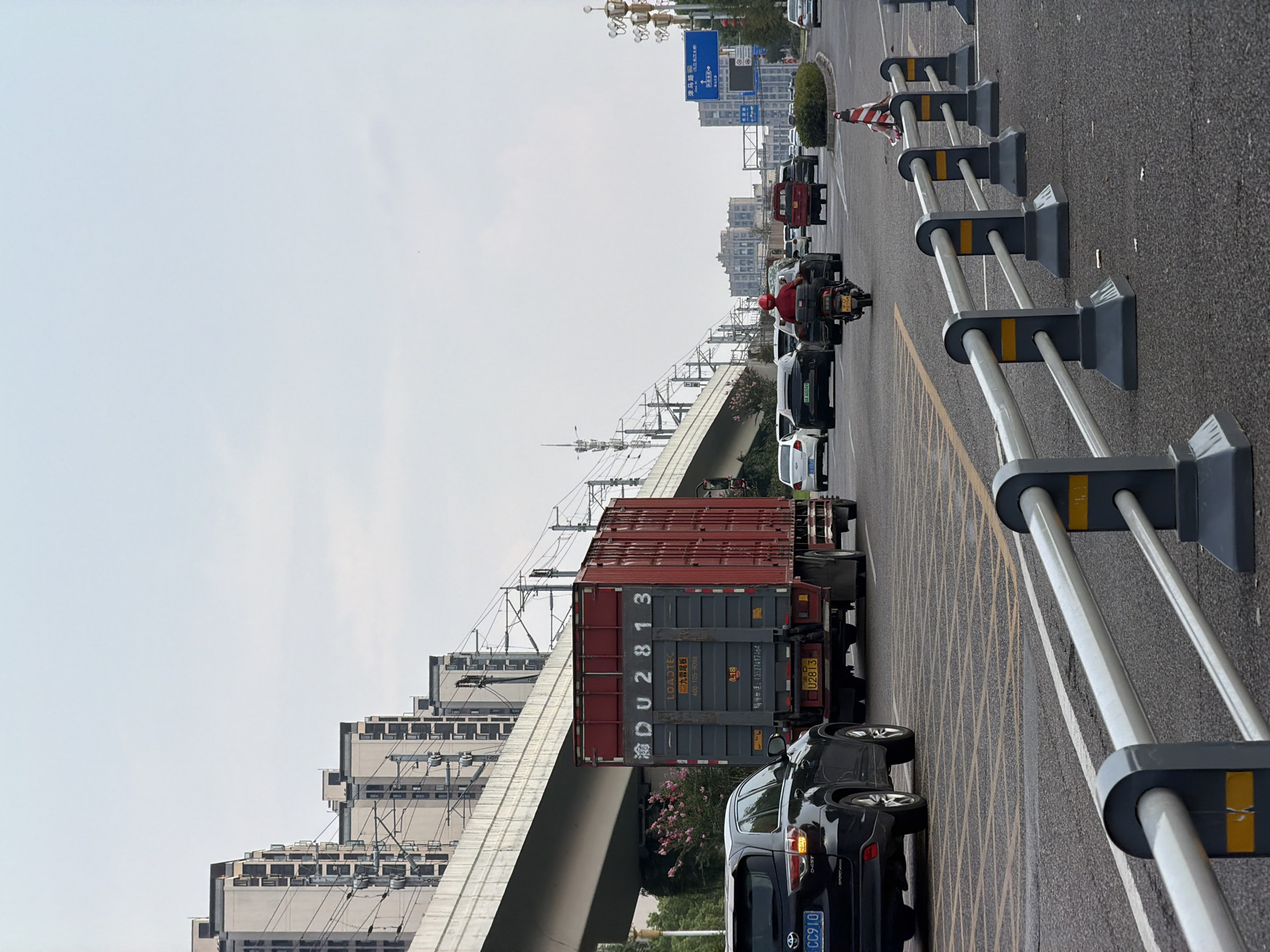 A traffic scene is depicted in Jiangjin, China, with a prominent overpass and apartment buildings in the background. In the foreground, a multi-lane asphalt road is observed with various vehicles. A black SUV with license plate "CC910" is in the left lane, facing away from the camera. To its right, a large red and gray container truck with license plate "U2813" is moving forward. A motorcycle with a rider wearing a red helmet is also in motion on the road, positioned behind the truck. In the middle distance, other cars and a red truck are visible, along with traffic cones and barrier fencing. The sky is overcast.