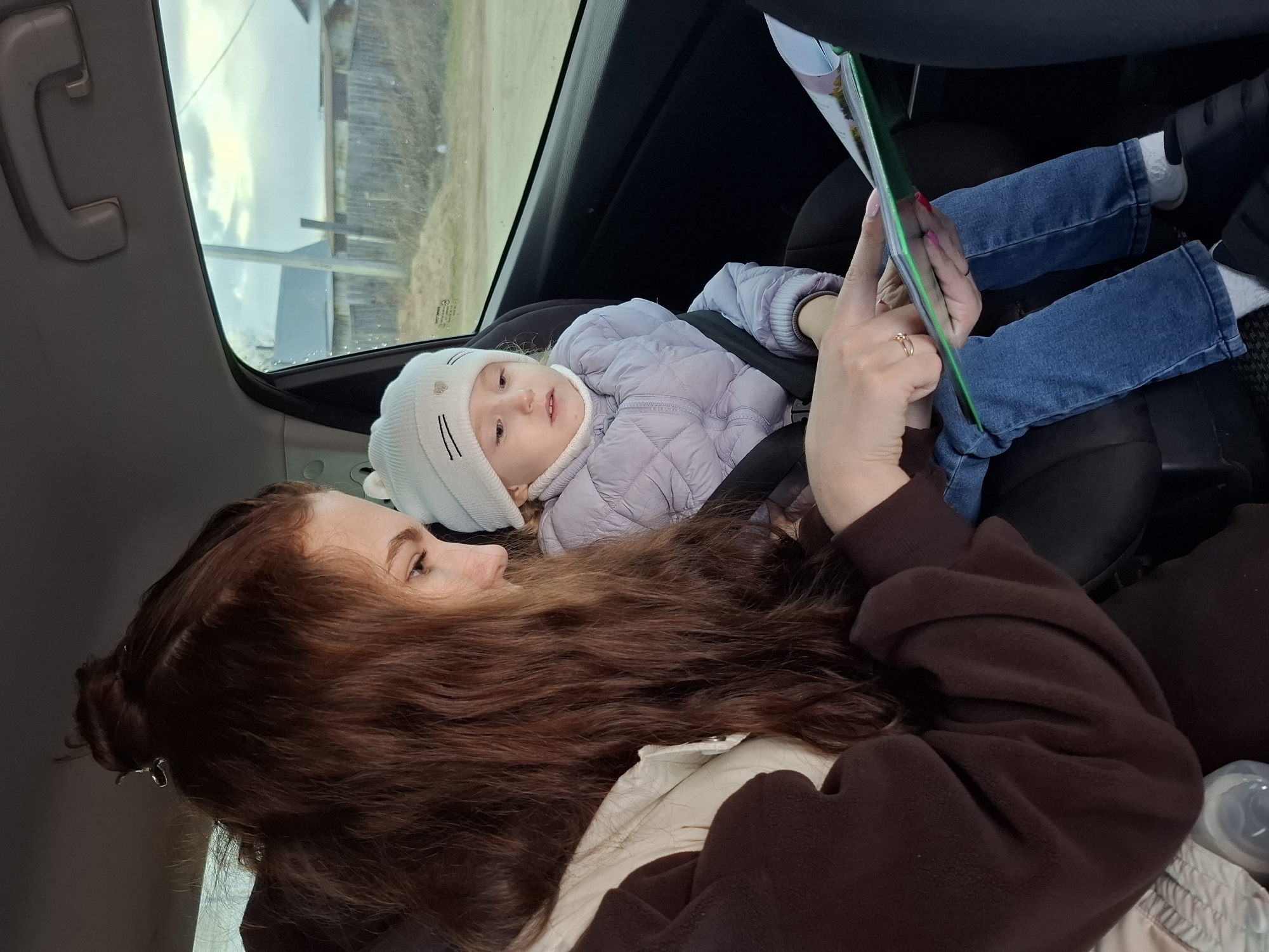 Inside a car, a woman with long, wavy reddish-brown hair, partially tied back, looks affectionately at a young child seated in a car seat. The woman is wearing a dark brown fleece top and a cream-colored layer underneath. She holds a green book or tablet, seemingly engaging the child with its contents.
The child, wearing a light purple quilted jacket, blue jeans, white socks, and white shoes, is secured in a black car seat. They also wear a white beanie with subtle cat ear details and heart-shaped embellishments. The child's expression is focused, looking towards the object the woman is holding.
The setting is the back seat of a car, indicated by the headliner with a gray grab handle and the visible window. Through the car window, an overcast sky is visible above a landscape featuring a wooden fence and some distant structures, suggesting a rural or semi-rural area, consistent with the provided location context of Гаревая, Russia. The time appears to be daytime due to the natural light filtering through the window. In the bottom right corner, a baby bottle is partially visible. A small, unreadable sticker with text is present on the car window glass. The woman wears a simple gold ring on her right hand.