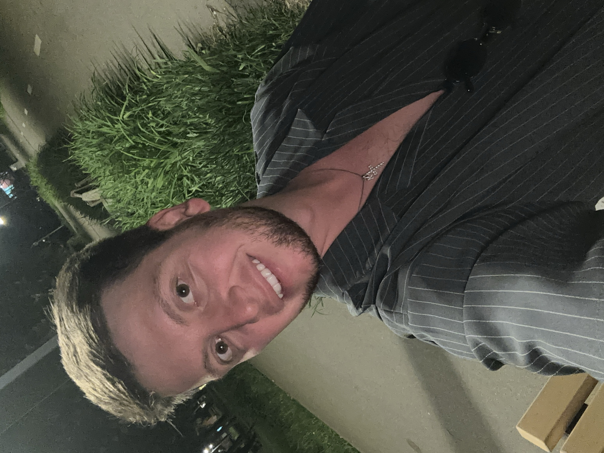 A man is shown in a medium shot, turned to face the viewer. He has short, light brown hair and a stubbled beard, and he is smiling. The man is wearing a gray pinstripe shirt or jacket with a low neckline and a delicate chain necklace. Sunglasses are perched on the upper portion of his garment. Behind him is a patch of green foliage and a paved area, suggestive of an urban setting. The overall illumination is subdued, indicating either nighttime or low lighting conditions. Moscow, Russia cannot be independently confirmed from this image.