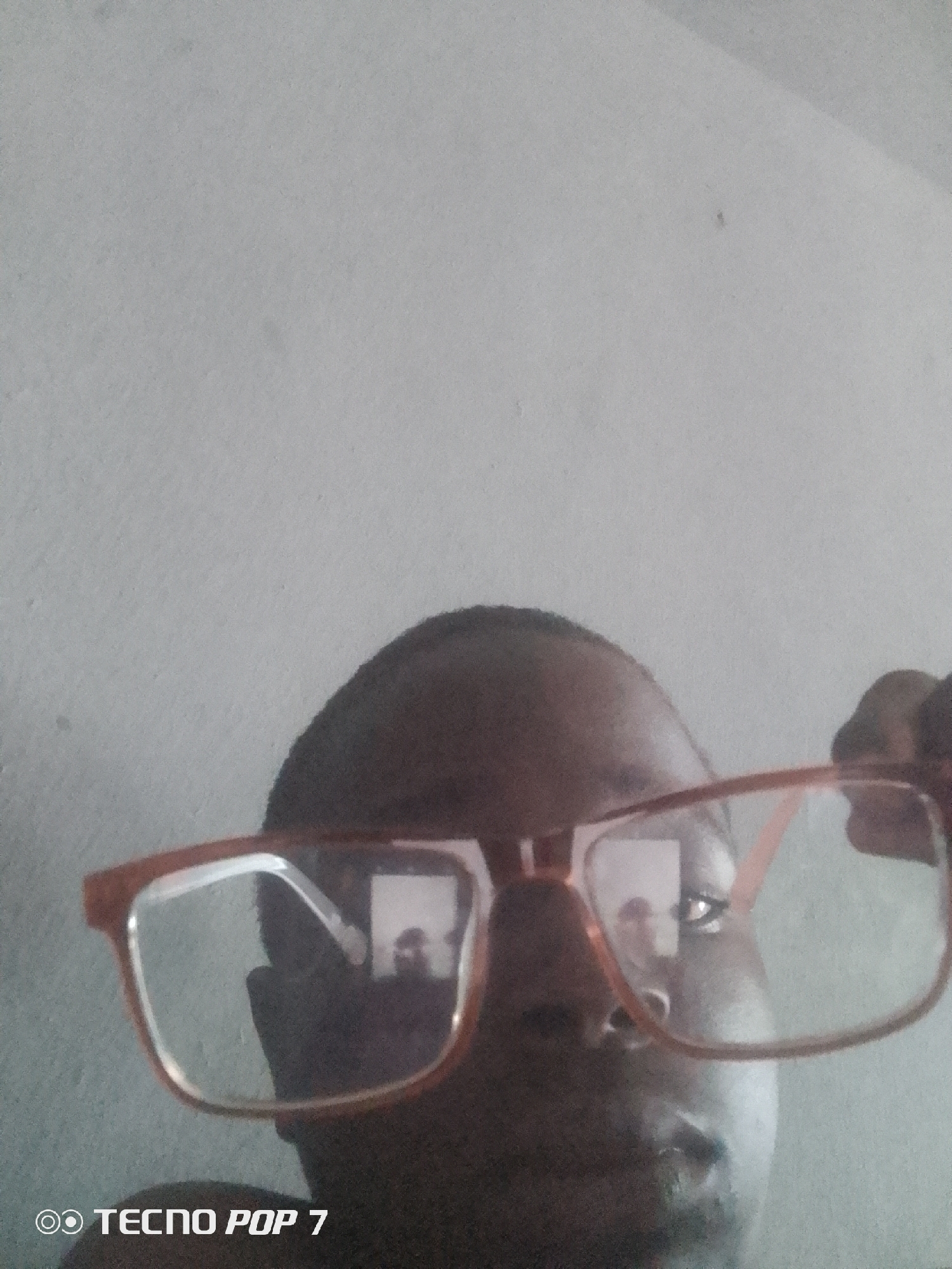 A medium shot shows a person holding a pair of orange-rimmed glasses in front of their face. The person's head is mostly obscured by the glasses, but their forehead, eyes, and nose are visible. The glasses are large and square-shaped with clear lenses. The person's eyes are looking directly at the camera through the lenses. The background is a plain, light gray wall. The bottom left corner of the image contains the text "TECNO POP 7" in white. The image appears to have been taken indoors in Ipetumodu, Nigeria.