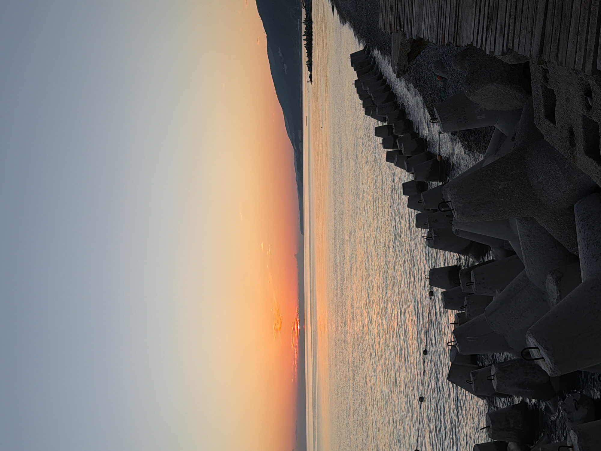 This image captures a serene coastal scene at sunrise in Sochi, Russia. The sky transitions from a soft, pale blue on the left to warm hues of orange and pink on the right, indicating the early morning light. A distinct, dark mountain range forms the horizon on the far left, partially silhouetted against the brightening sky.
The foreground is dominated by a body of water, likely the Black Sea, with gentle ripples reflecting the dawn colors. Along the right edge of the water, a breakwater made of large, concrete tetrapods is clearly visible. These geometric structures are designed to absorb wave energy and protect the coastline. Beyond the tetrapods, a wooden walkway or pier stretches into the distance, providing a stark contrast in texture and color to the natural elements.
The overall mood is peaceful and tranquil, evoking a sense of calm and the quiet beauty of a new day beginning. The time of day is definitively sunrise. There are no people visible in the frame. The location is cued by the mention of Sochi, Russia, and the characteristic coastal landscape with breakwater structures. No visible text is present in the image.
