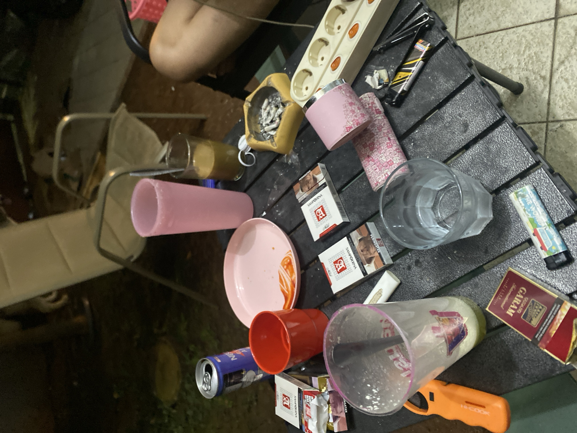 The image depicts a low-angle view of a dark, outdoor setting, likely at night. A black, foldable table is covered with various objects, suggesting a casual gathering. These include: a blue and silver can, a red plastic cup, a pink plate with what appears to be sauce residue, two packs of cigarettes (one white with red lettering, the other red with gold lettering), a clear plastic cup with a straw and residual liquid, another clear glass with liquid, a pink tumbler, a yellow ashtray filled with cigarette butts, a power strip with multiple sockets, an orange lighter, and a lighter with a yellow and black design. In the background, the legs of two chairs are visible. The ground appears to be a mix of dark soil and a paved area with light-colored tiles. The overall impression is one of a relaxed, informal social environment. The location is identified as Dukuh, Indonesia.