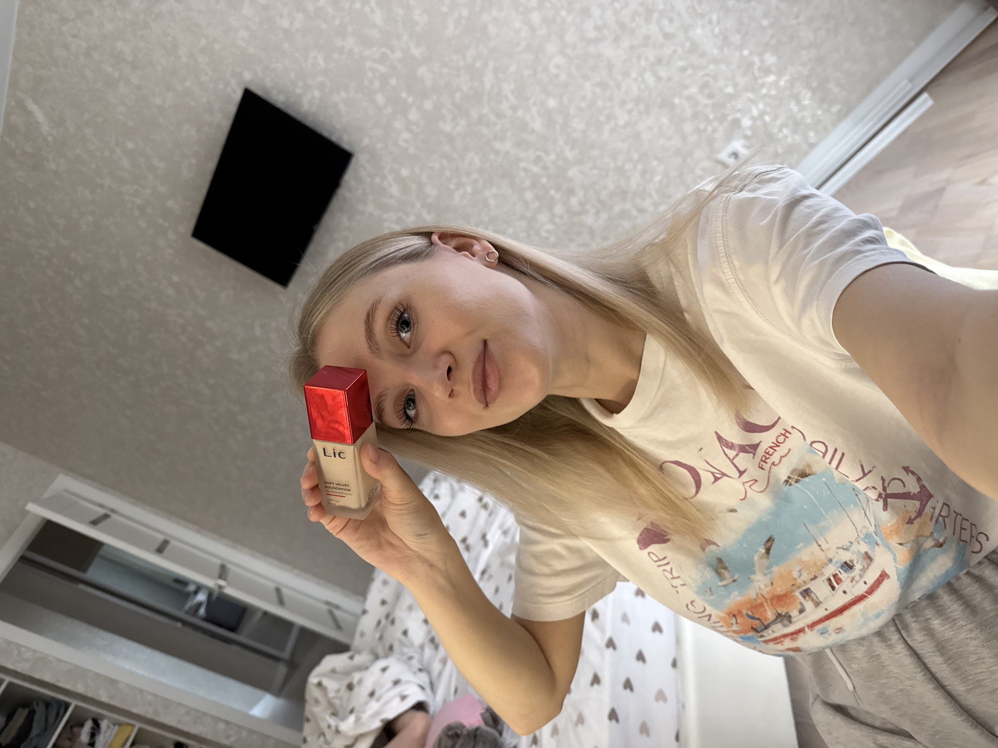 A young woman is holding a bottle of "Lic Soft Velvet Foundation" makeup in her left hand. She is positioned inside what appears to be a bedroom or dressing area. To the left, a wardrobe or closet with multiple doors and compartments is visible. The ceiling is textured and patterned. A black, square object is affixed to the wall, potentially a light fixture or a decorative panel. The woman is wearing a white t-shirt with a nautical design and gray sweatpants. The specific location in the city of Izhevsk, Russia, cannot be determined from the available visual information.