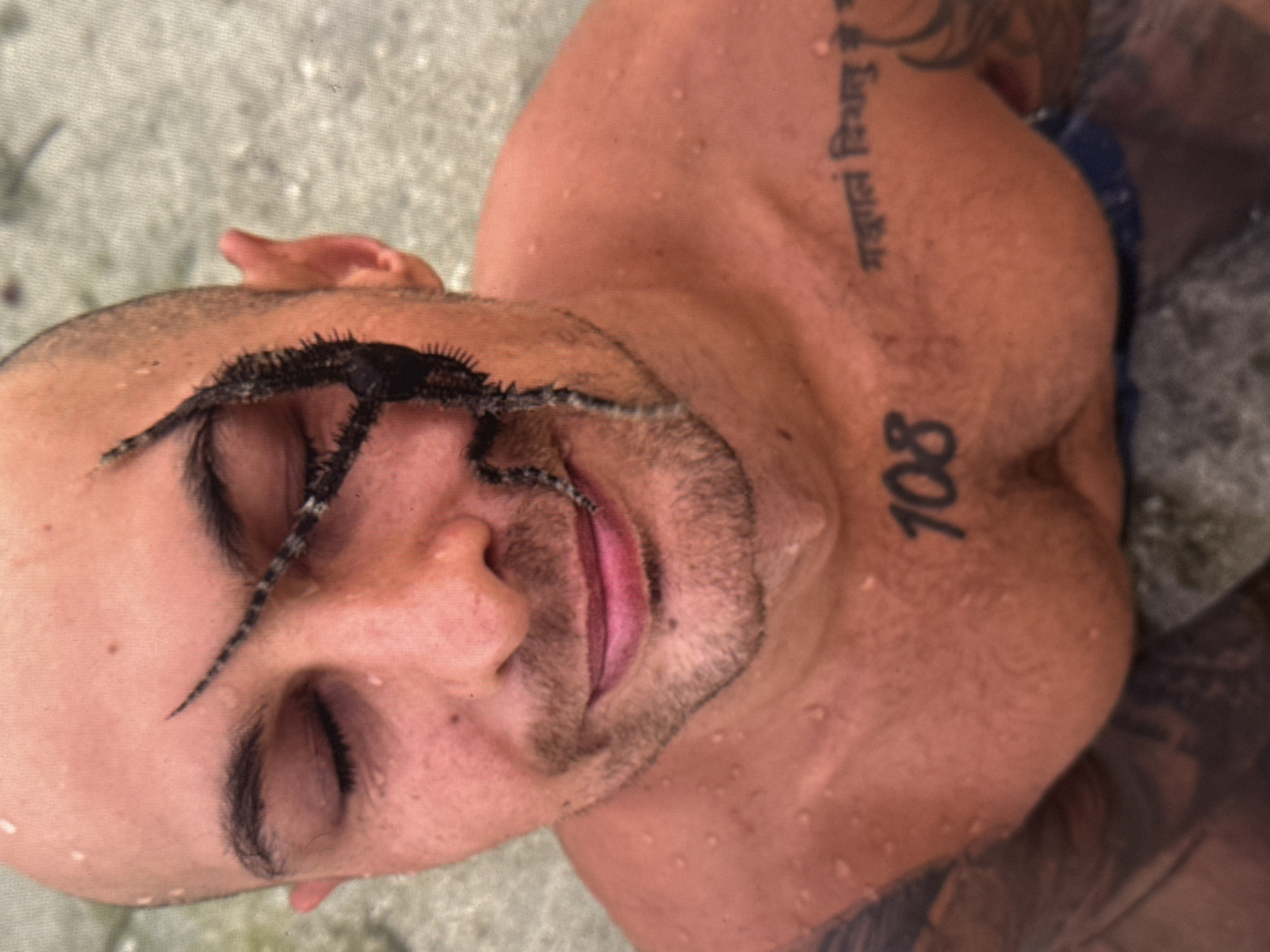 A close-up, high-angle shot shows a man with a shaved head and a sea creature on his face. The man's eyes are closed, and he has a slight smile. The sea creature, which appears to be a brittle star, is black and has multiple arms that extend across his face. The man's skin is wet, with water droplets visible on his forehead and chest. He has tattoos on his chest and left arm. The background is blurry and appears to be water or sand. The image is likely taken in Kuta Selatan, Indonesia, given the man's tattoos which appear to be in Sanskrit, a language common in Bali, Indonesia. The number '108' is tattooed on the man's chest.