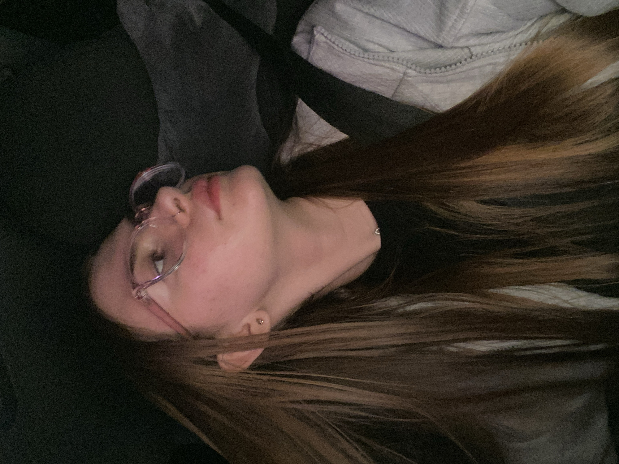 A young woman with long, brown hair and glasses is looking upwards and to the right. She is wearing a black t-shirt and a light-colored jacket, and a seatbelt is visible across her chest. A small earring is visible in her left ear, and a nose ring is also present. The background is dark and out of focus, suggesting she may be in a vehicle at night. The provided crops offer close-ups of her face, hair, and clothing, highlighting the details of her features and attire. The image does not provide any information about the city of Дьяково, Russia.