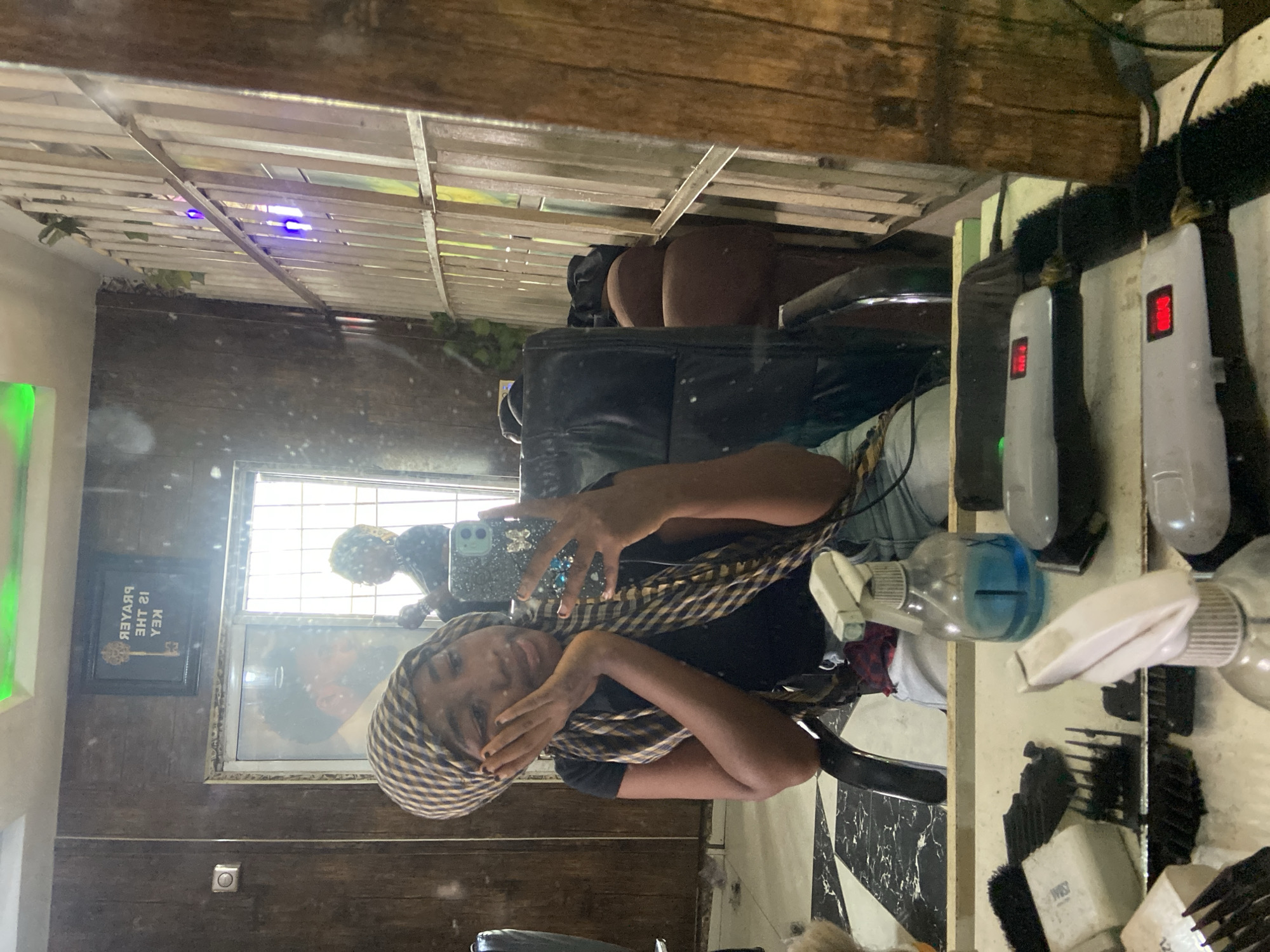 The image is a mirror selfie taken in what appears to be a barbershop or hair salon, with the reflection capturing the subject and the interior of the establishment. The main subject is a dark-skinned individual, likely a woman, wearing a black top and a checkered headwrap (brown/beige and black/dark blue). She is holding a smartphone with a decorated case (featuring rhinestones or gems) in her right hand, capturing her reflection, while her left hand covers part of her face. She has dark nail polish and displays a slight smile, suggesting a relaxed or happy demeanor.
The setting features dark wooden paneling on the walls and a slatted wooden structure overhead, possibly a decorative ceiling or partition. In the background, another person with dark, possibly curly hair, is faintly visible through a window's reflection. On a counter to the right, various salon tools are neatly arranged, including multiple barber clippers or trimmers with red digital displays, several spray bottles (one with blue liquid, others clear), and brushes and combs. A black salon chair is also partially visible.
Notable details include purple/blue lights emanating from the slatted ceiling structure and a bright green light source on the far left. The floor visible on the bottom right has a black and white checkered pattern. On the left wall, a framed picture is reflected, clearly displaying the mirrored text "PRAYER IS THE KEY." The overall activity is a mirror selfie being taken in a professional hair care environment in Lokoja, Nigeria.