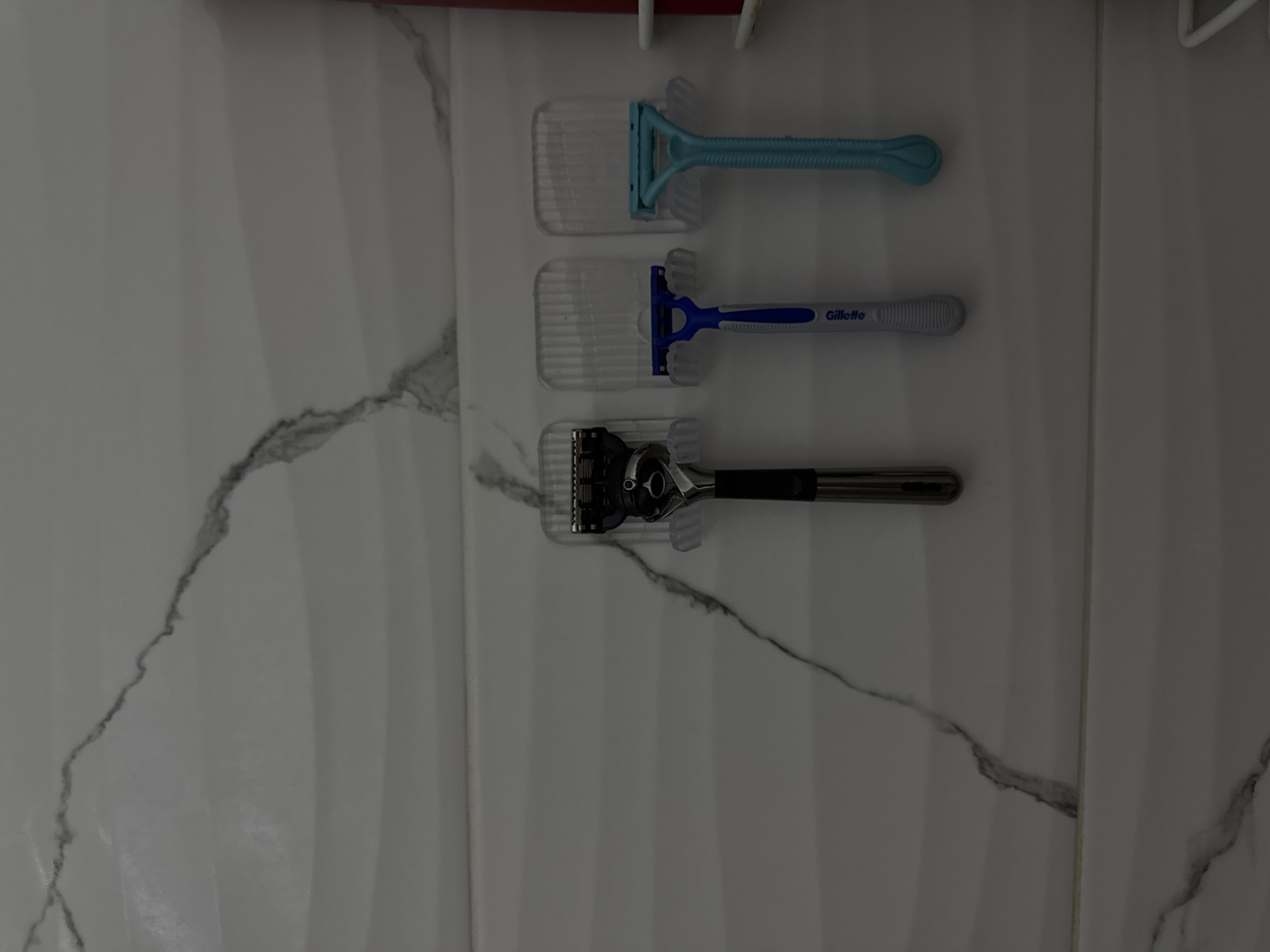 A vertically oriented photograph displays three razors mounted on a white, marble-patterned wall. The razors are arranged vertically, each in its own clear plastic holder. The top razor is a light blue disposable type. The middle razor is a blue and white Gillette brand. The bottom razor has a black handle and a silver head with multiple blades. The wall appears to be in a bathroom or similar space, given the presence of razors. The photograph does not indicate any information about the location in Sochi, Russia. The scene includes objects, razors in the razor holders and the environment, the marble pattern wall.