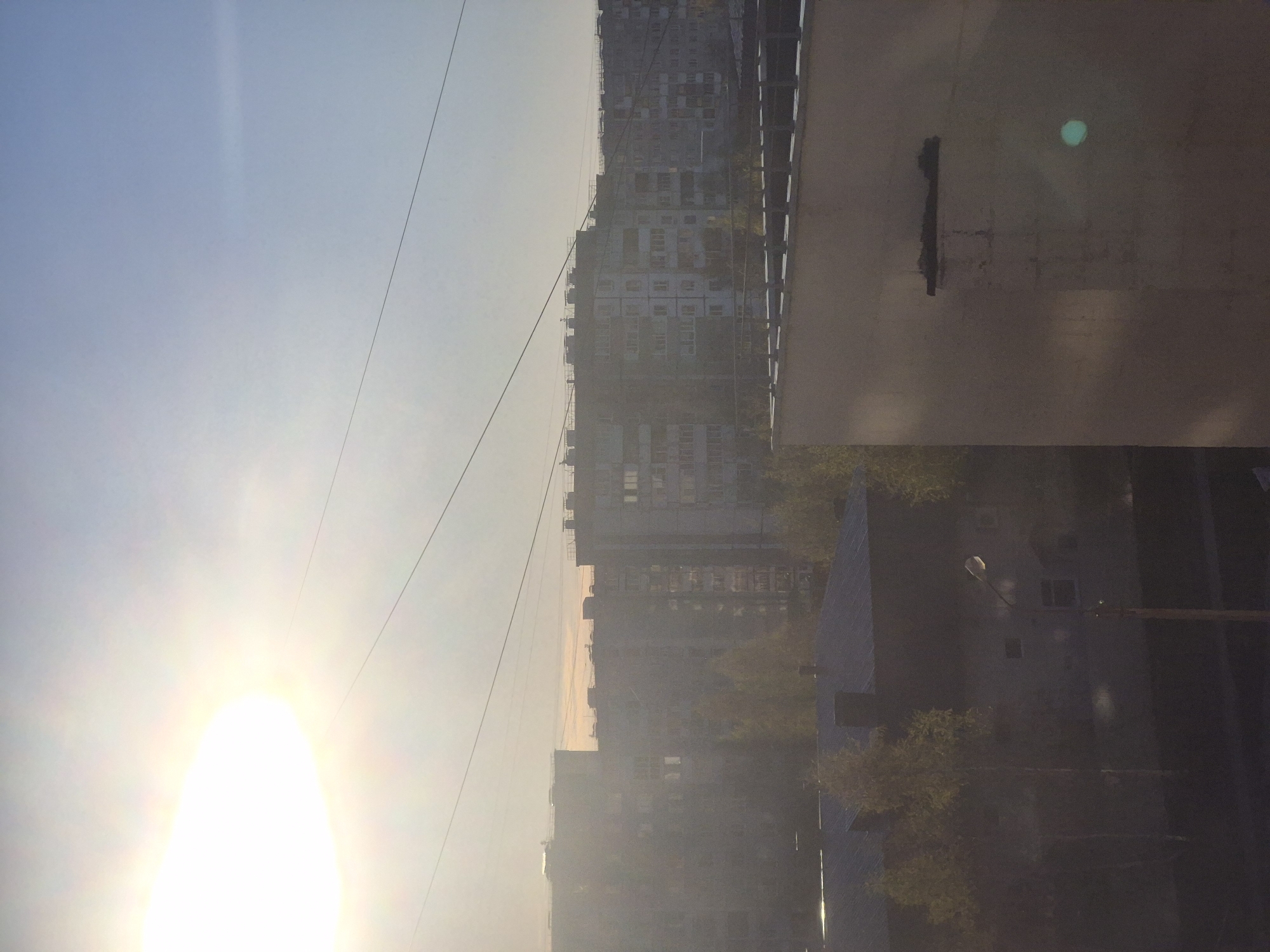 The image shows a view of a cityscape in Tobolsk, Russia. The sun is positioned in the sky, and power lines stretch across the view. Tall apartment buildings are visible in the background. Part of a building juts into the frame on the right side of the picture. The environment appears hazy, possibly due to fog or air pollution. A street lamp can be seen. There is no visible evidence of people.