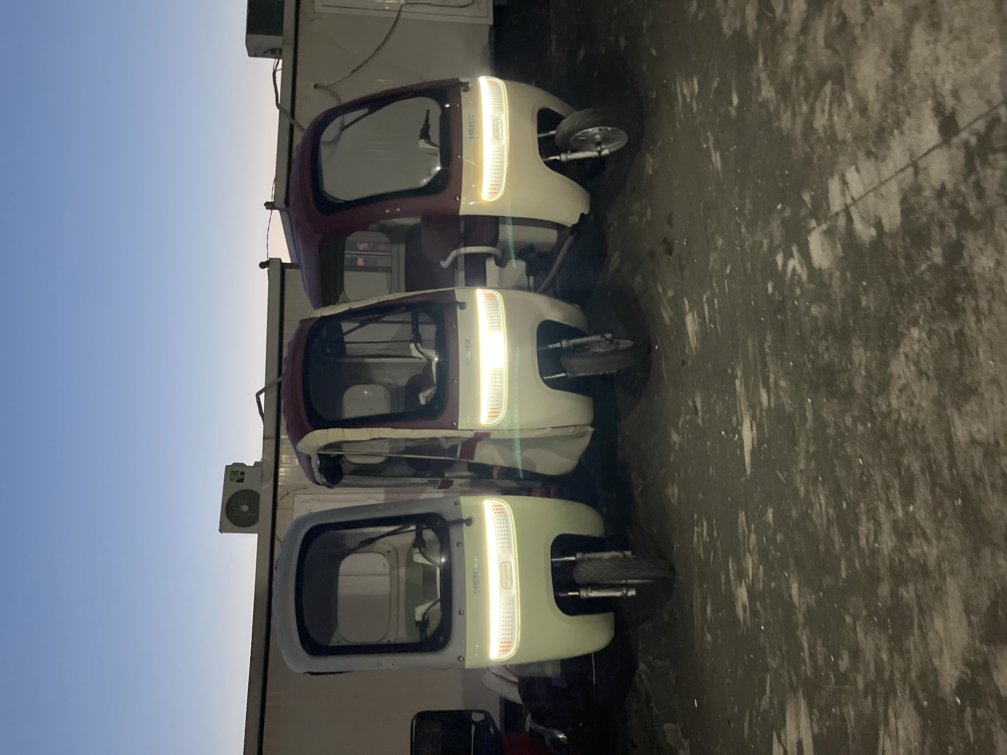 The image shows three enclosed three-wheeled vehicles parked on a dirt or gravel surface. The vehicles are light-colored with darker colored roofs. They have headlights that are illuminated. In the background, there is a long, light-colored building with an air conditioning unit visible on the roof. The sky is a gradient of blue and gray, suggesting it is either early morning or late evening. The overall scene appears to be an outdoor storage or parking area. The location is specified as Ureki, Georgia.