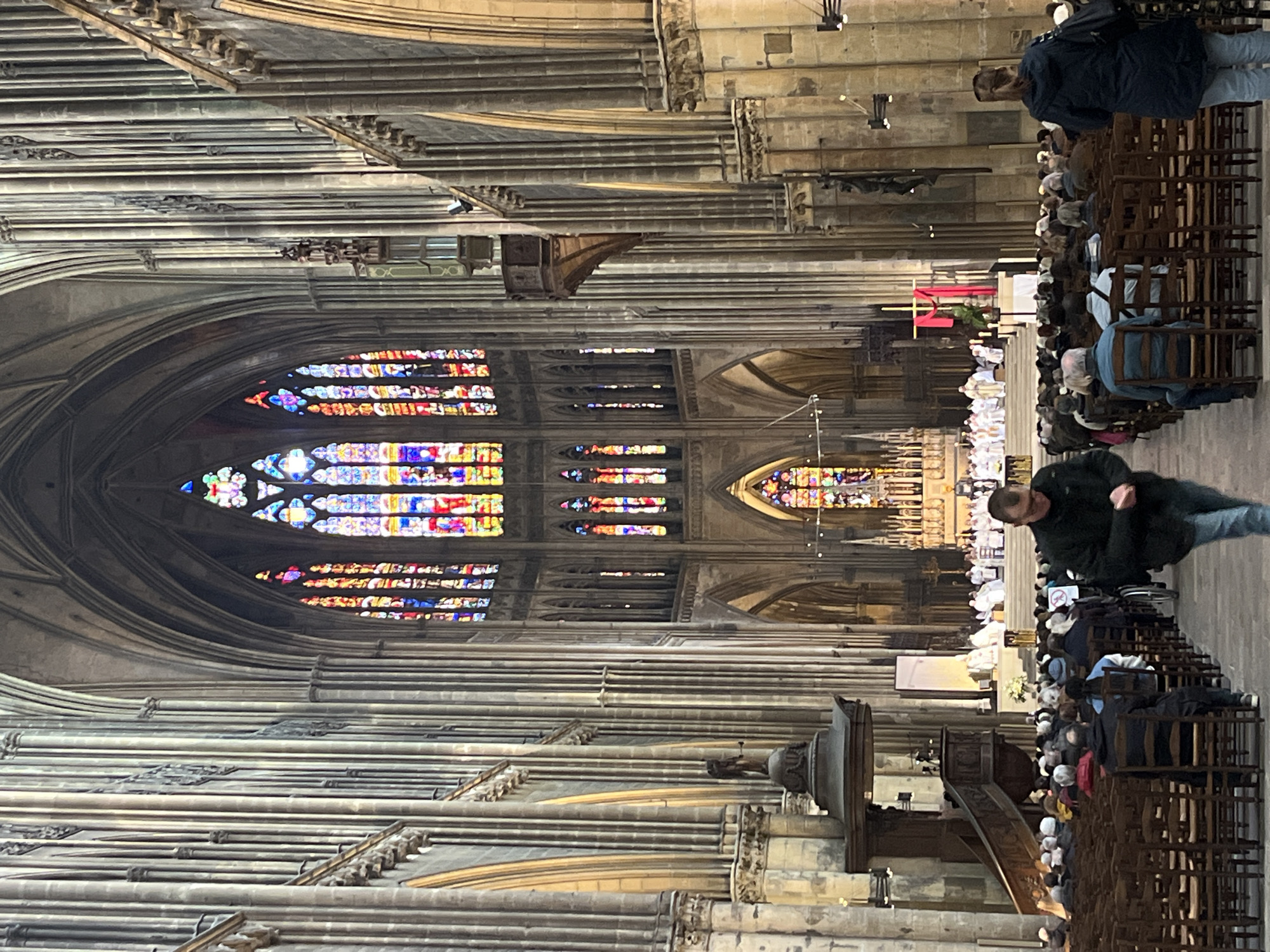 The image captures the interior of Metz Cathedral in Metz, France. The architecture is predominantly Gothic, with high vaulted ceilings, ribbed vaults, and stained glass windows. The windows display intricate patterns and vibrant colors. People are seated in rows of wooden chairs, likely attending a religious service or event. In the center of the image, near the altar, a procession of people in white robes can be seen. Two people are walking on the aisle, one wearing a black jacket and one wearing a blue jacket. A wooden pulpit stands on one side. The environment is well-lit, with natural light streaming through the stained glass windows.