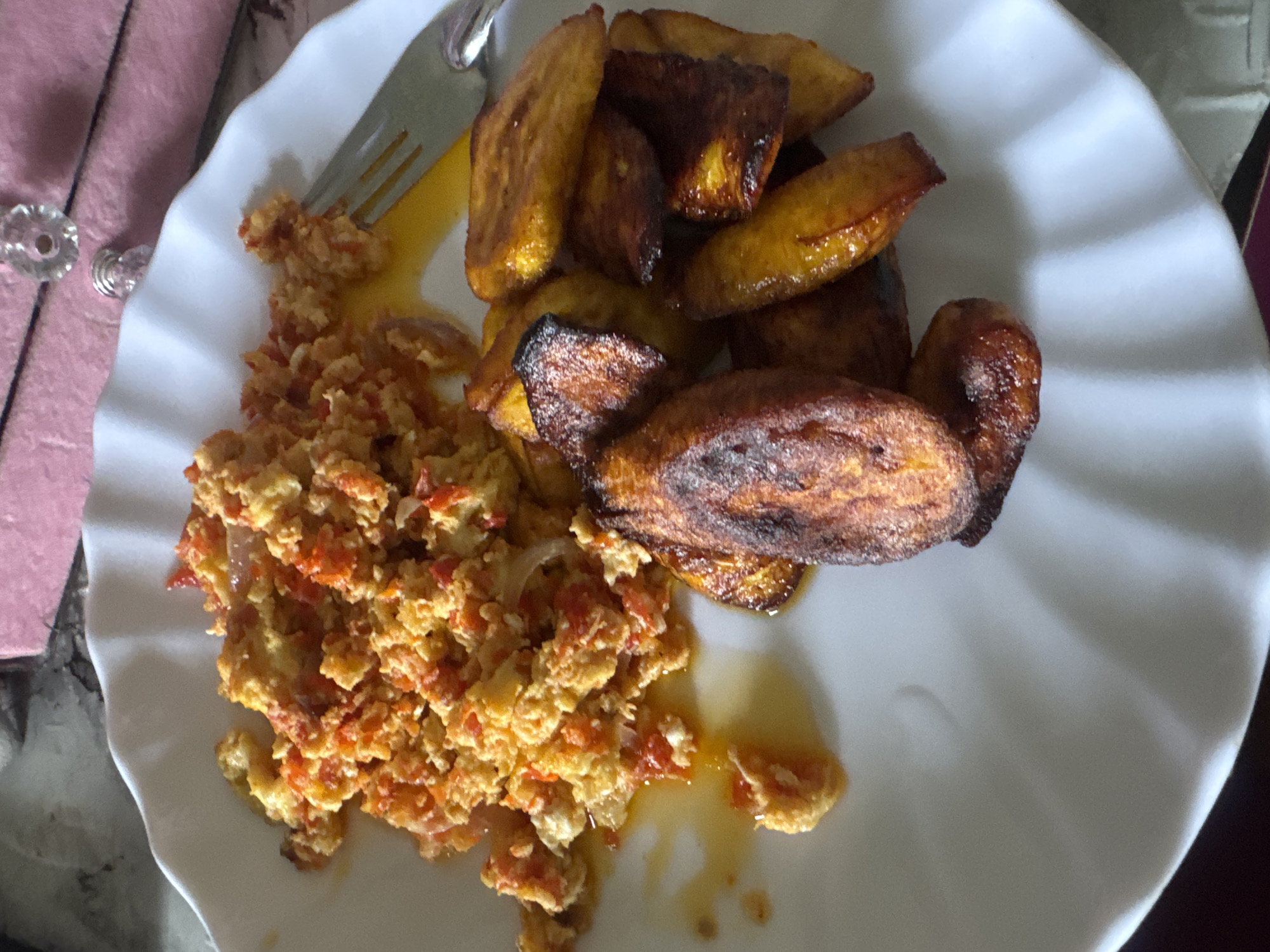 An overhead, close-up shot captures a meal served on a white, fluted plate, likely in Gbongan, Nigeria. The plate holds two main components: a generous portion of scrambled eggs mixed with finely diced red ingredients and some lighter pieces, presumably tomatoes, peppers, and onions; and a pile of deeply fried plantain slices (dodo).
The scrambled eggs exhibit a vibrant orange-yellow hue, with the red vegetable pieces integrated throughout, suggesting a flavorful preparation. A sheen of oil or sauce surrounds the base of the eggs, indicating they are freshly cooked or have been dressed. To the right of the plate, the fried plantains are golden to dark brown, some pieces showing well-caramelized edges, reflecting thorough frying.
A silver-colored metal fork is partially visible on the upper left side of the plate, with its tines resting near the egg mixture and some oil. In the blurred background, a textured, soft pink or purple fabric, possibly a tablecloth or napkin, is visible. Adjacent to it, a decorative, clear, faceted knob, likely part of furniture or decor, adds a domestic touch. The setting appears to be indoors, potentially a home kitchen or dining area, and the meal itself is typical Nigerian fare. No text is visible in the image.