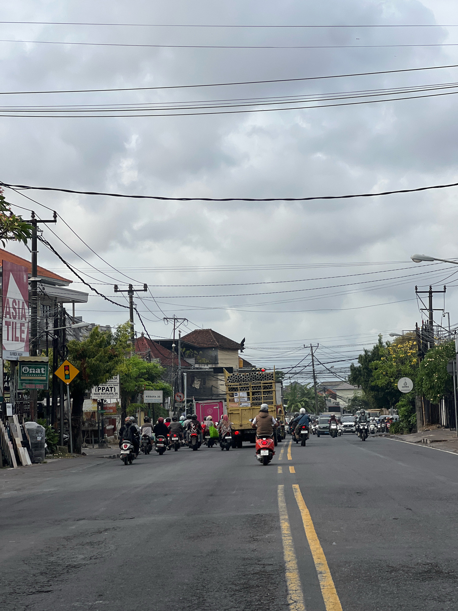 The image captures a street scene in Denpasar, Indonesia, under an overcast sky. The foreground features a paved road with yellow lane dividers, showing some wear and tear. A procession of motorcycles and a truck dominate the roadway, indicating moderate traffic. Several individuals, mostly on motorcycles, are visible, some wearing helmets. Buildings line the street, featuring commercial signage including "ASIA TILE" and "Onat Hardware." Utility poles with a network of electrical wires crisscross the sky. Trees and foliage are interspersed among the buildings, adding greenery to the urban environment. The overall atmosphere suggests a typical day in a bustling Indonesian city.