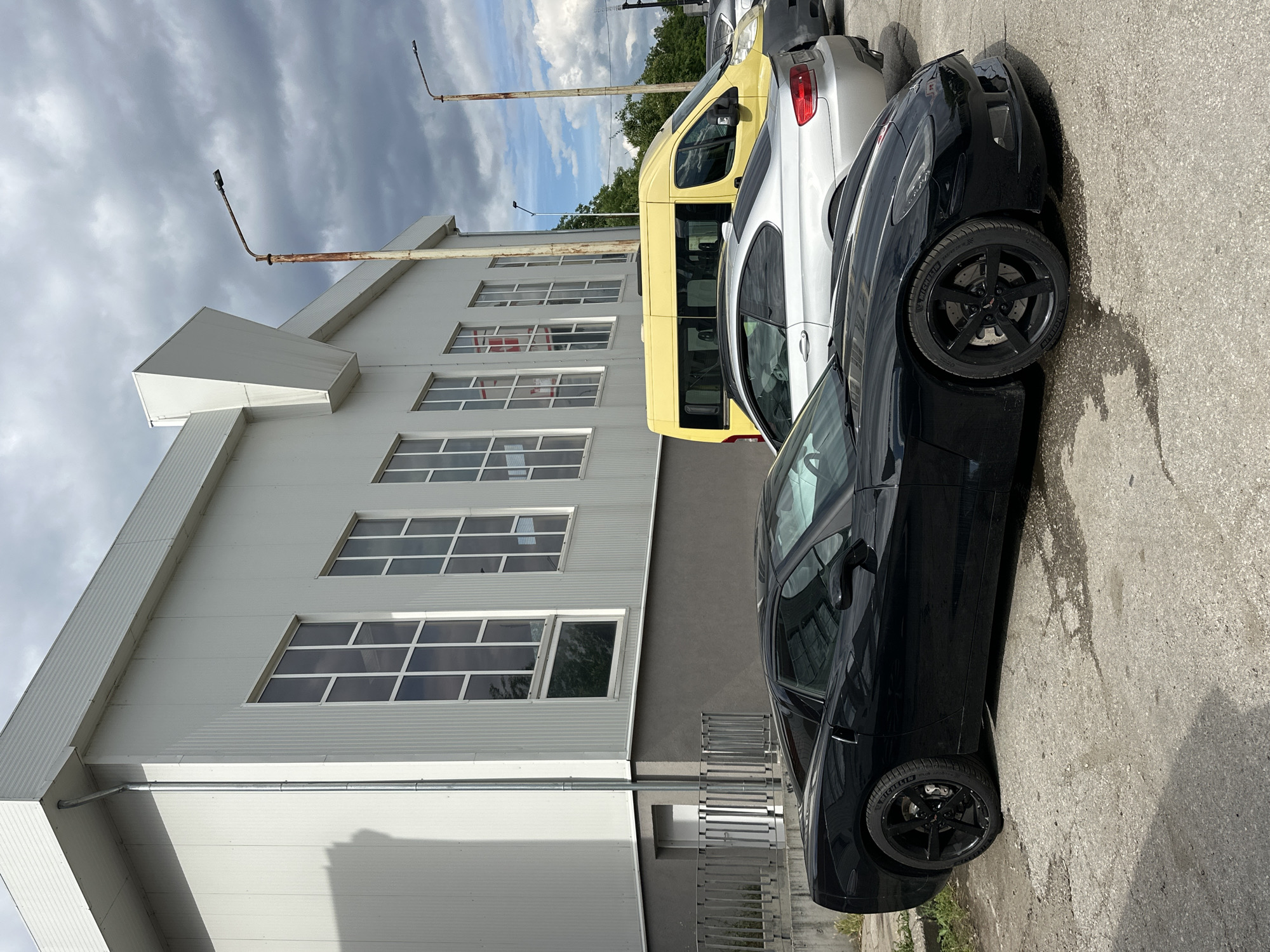 A black Chevrolet Corvette is parked on a concrete lot in front of a white industrial-style building with numerous large windows. To the right of the Corvette, a silver BMW coupe is parked, followed by a yellow van. The building has a grey base and a metal corrugated roof. Two rusty streetlights stand near the building. The sky is overcast with dramatic clouds. The image appears to have been taken in Veliko Tarnovo, Bulgaria, as suggested by a tag on an external dataset.