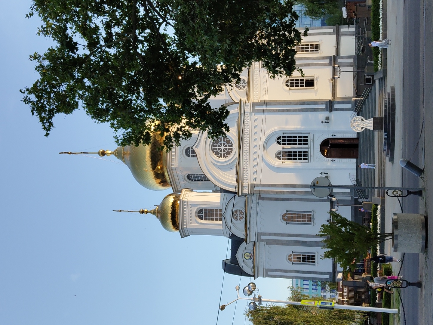 This is a full, frontal, eye-level shot of the Catherine Cathedral in Krasnodar, Russia. The sun is setting, casting a warm glow on the white stone facade of the cathedral. The cathedral has a large, ornate entrance with wide stairs leading up to it. Two golden domes, topped with crosses, rise above the main structure. A large, leafy tree obscures the right side of the cathedral. In front of the cathedral, there is a public square with a fountain and a sculpture. People are seen walking and standing in the square, and a few cars are visible in the background. The sky is clear and blue.
