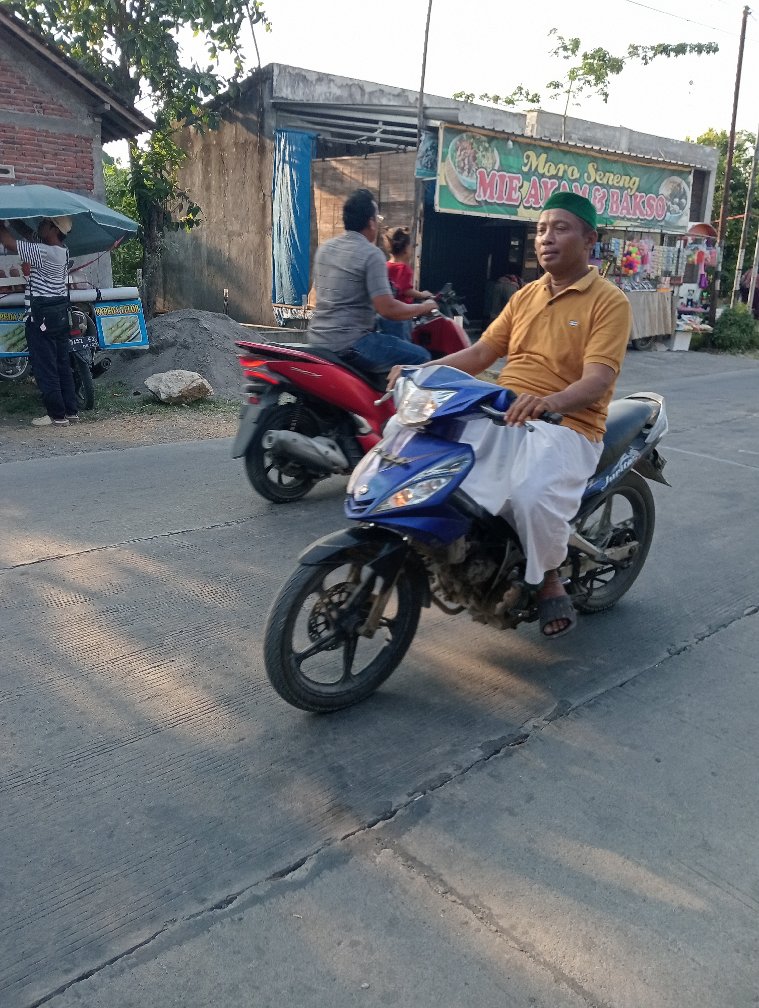 In Tanjunganyar, Indonesia, a man wearing a yellow polo shirt and a green cap rides a blue motorcycle. He is wearing white pants and dark sandals. Behind him, another motorcycle, red in color, is also in motion, with its rider wearing a grey shirt and blue jeans. In the background, a food stall displays a sign that reads "Moro Seneng MIE AYAM & BAKSO." Adjacent to the stall, a vendor stands near a cart under a green umbrella. The scene takes place on a paved road with visible cracks, bathed in the warm light of what appears to be late afternoon or early morning.
