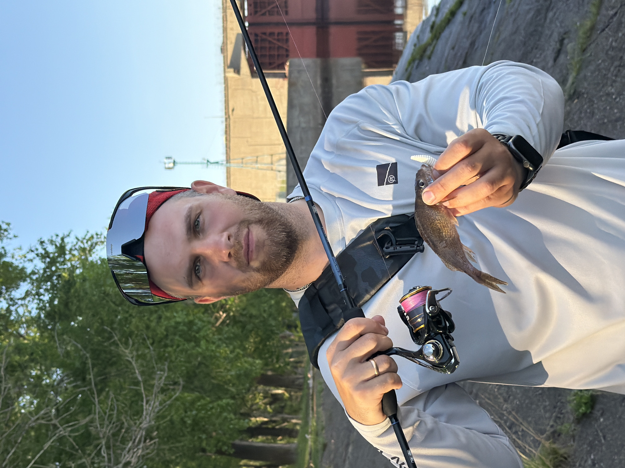A fair-skinned man with short hair and a beard is holding a small fish in his right hand. He is wearing sunglasses, a long-sleeved white shirt, and a black watch. He also has a black harness-like strap across his chest. In his left hand, he holds a fishing rod with a black and silver reel. The background includes a large concrete structure with red accents, and some trees. The image was taken in Chaikovsky, Russia.