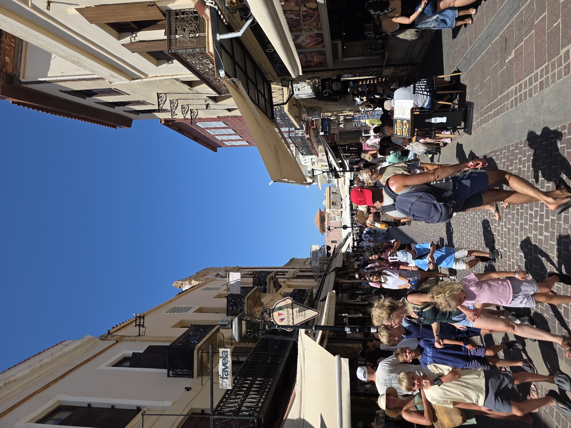 The image shows a bustling pedestrian street in Chania, Greece, likely in the Old Town. The street is paved with cobblestones and lined with multi-story buildings featuring traditional architecture, including balconies with wrought-iron railings and wooden shutters. Shops and eateries are visible on the ground floor, with awnings providing shade. A clear blue sky indicates fair weather.
Numerous people are walking along the street, dressed in casual summer attire. They appear to be tourists and locals enjoying the atmosphere. Some individuals are in small groups, while others are walking alone. The age range of the people visible varies, with families and children present.
Objects in the scene include street lamps, shop signs (one depicting an ice cream cone with Greek text), umbrellas, tables and chairs outside a cafe, and shopping bags. The overall impression is one of a vibrant, lively urban environment.