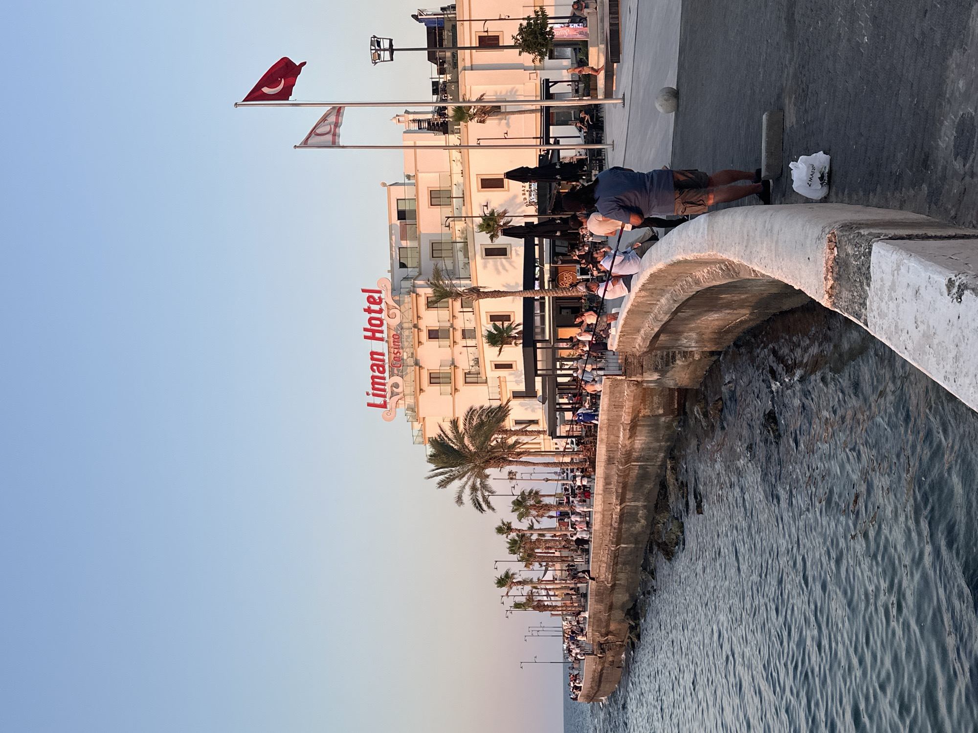 The image depicts a coastal promenade in Kyrenia, Northern Cyprus, with the Liman Hotel and Casino prominently featured. The setting is illuminated by the soft light of either sunrise or sunset, casting a warm glow on the scene. Several individuals are visible along the curved seawall, some engaged in conversation, others observing the sea or enjoying the ambiance. A man in the foreground is facing away from the camera, holding a fishing rod, suggesting a recreational activity. Further along the promenade, groups of people are seen dining or socializing at outdoor seating areas of establishments connected to the hotel. Palm trees are interspersed along the walkway, adding a tropical feel to the landscape. Two flags are prominently displayed near the hotel: one is the Turkish flag, and the other is the flag of the Turkish Republic of Northern Cyprus. The sea is calm, with gentle waves lapping against the stone embankment. A white plastic bag rests on the paved walkway in the foreground. The overall scene conveys a relaxed and lively atmosphere in a seaside resort town.