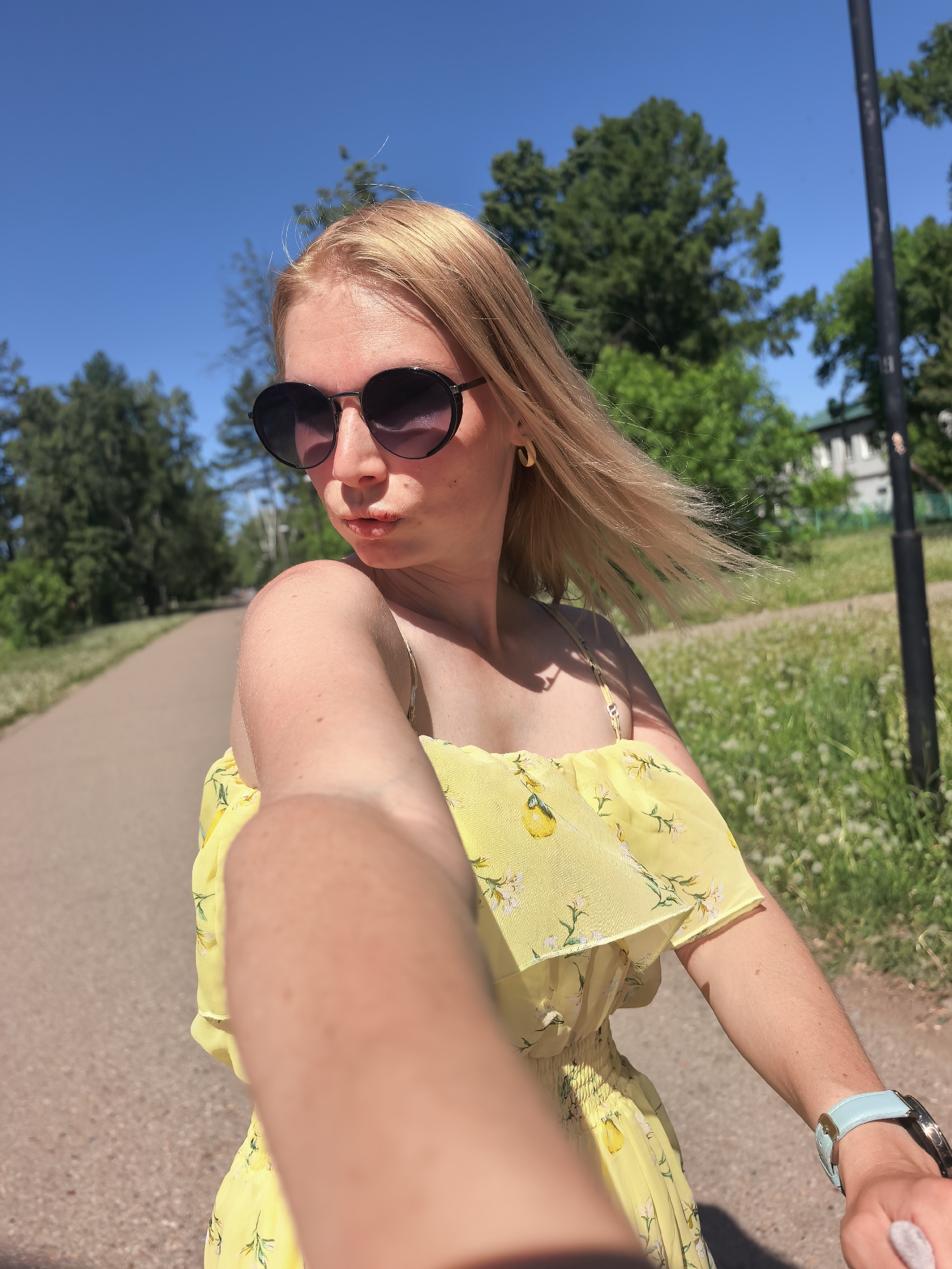 A blonde woman wearing a yellow, floral dress and round sunglasses is taking a selfie on a paved path in a park. She has gold hoop earrings. The sky is blue and sunny with green trees in the background. A black pole is to her right. The location is Sterlitamak, Russia, based on the presence of specific architectural features and the local flora visible in the background. The path is bordered by grass and wildflowers. The woman is holding her arm out towards the camera, obscuring some of her dress. A blue smartwatch is visible on her wrist.