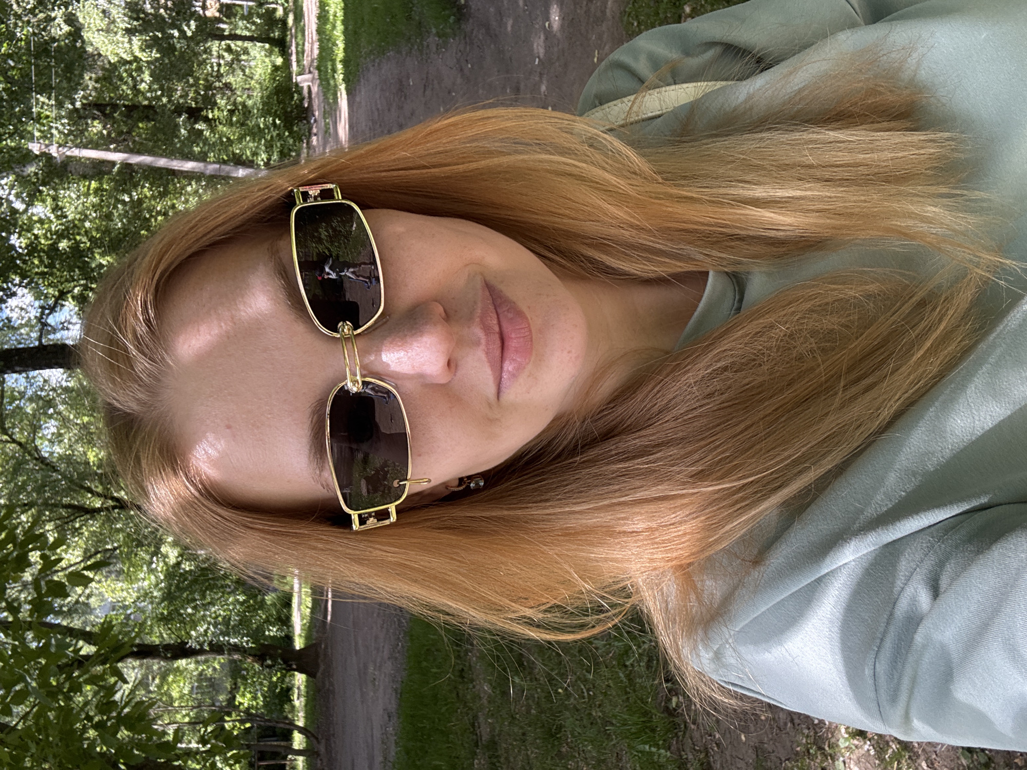 A close-up selfie shows a woman outdoors in a sunny, natural environment, likely in Rusanovo, Russia. The woman has long, reddish-blonde hair and is wearing a light green or mint-colored hoodie. She wears distinctive gold-framed sunglasses with dark lenses, which obscure her eyes. A notable reflection of a figure or object on a dirt path can be seen in the left sunglass lens, hinting at the surroundings behind the person taking the photo. Her lips are slightly parted, and her expression appears neutral to subtly content. The background features lush green trees and foliage, with a dirt path or road visible winding through the greenery, suggesting a park or forest setting. The bright natural light indicates it is daytime. No visible text is present in the image.