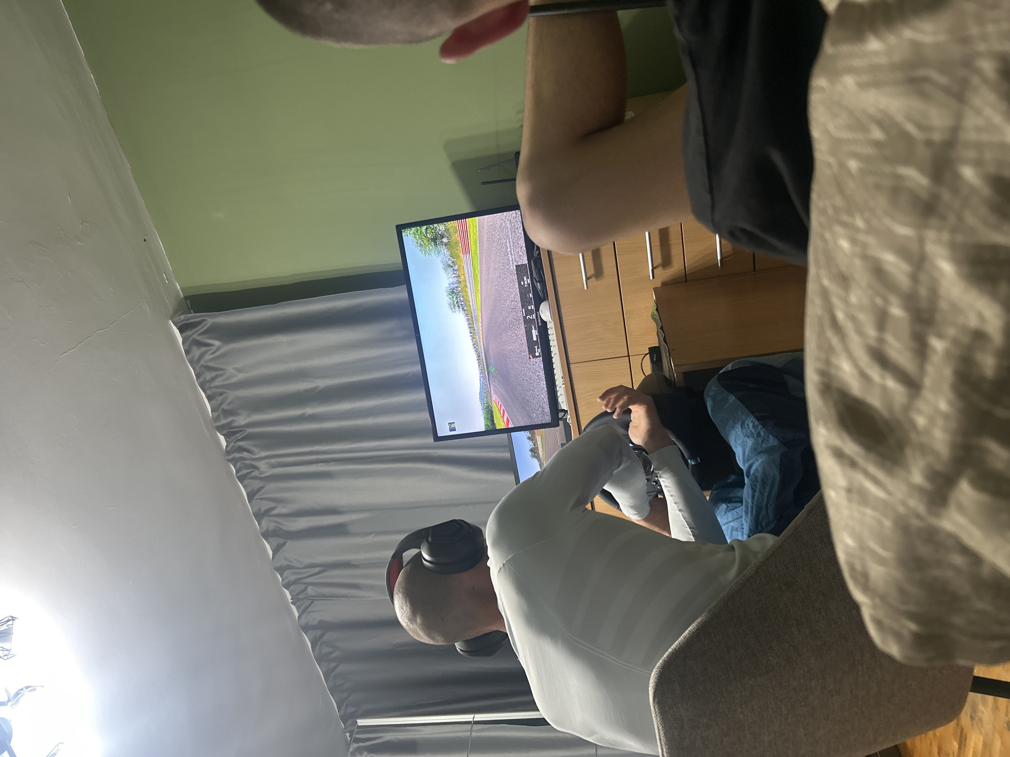 A man wearing headphones and a white long-sleeve shirt is playing a driving video game on a screen. He is using a steering wheel. A second person is partially visible in the top of the frame. The room is lit dimly. There are gray curtains on the left side. There are light wood cabinets to the right of the screen. The location is likely an apartment in Burgas, Bulgaria, based on the context.
