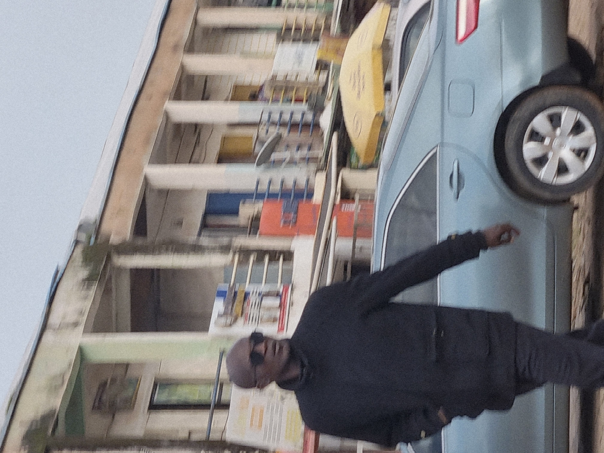The image shows a person wearing sunglasses and a dark long-sleeved shirt, with their arm raised. They are standing next to a light blue car. In the background, there is a multi-story building with a light-colored facade and balconies. The building appears to be under construction or in disrepair, with exposed concrete and some scattered debris. There are also some yellow and red elements visible on the building's exterior, possibly signs or construction materials. The sky is overcast, suggesting it might be daytime with cloudy weather. The overall scene appears to be an urban setting in Jos, Nigeria. No visible text is discernible in the image.