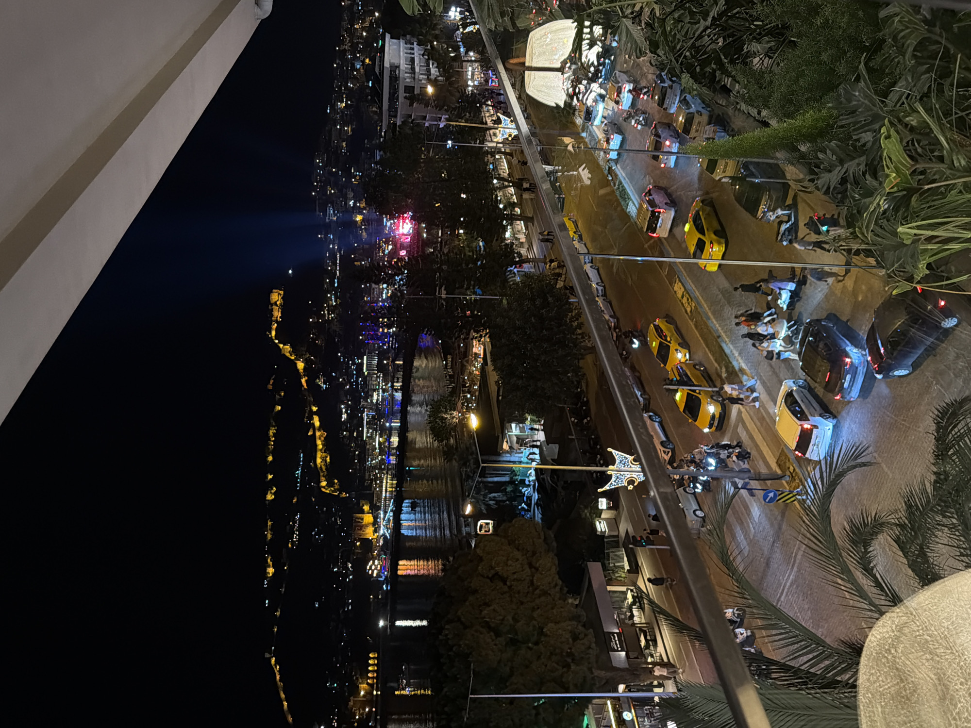 The image captures a nighttime view of Alanya, Turkey, from an elevated perspective, likely a balcony. The city's lights illuminate the landscape, creating a vibrant scene. To the left, a dark area contrasts with the brightly lit cityscape. A body of water, possibly a river or harbor, reflects the lights. In the middle ground, a street is visible, populated with vehicles, including several yellow taxis. Pedestrians are also seen on the sidewalks, some near parked motorcycles. Decorative streetlights and greenery, like palm trees, add to the urban environment.