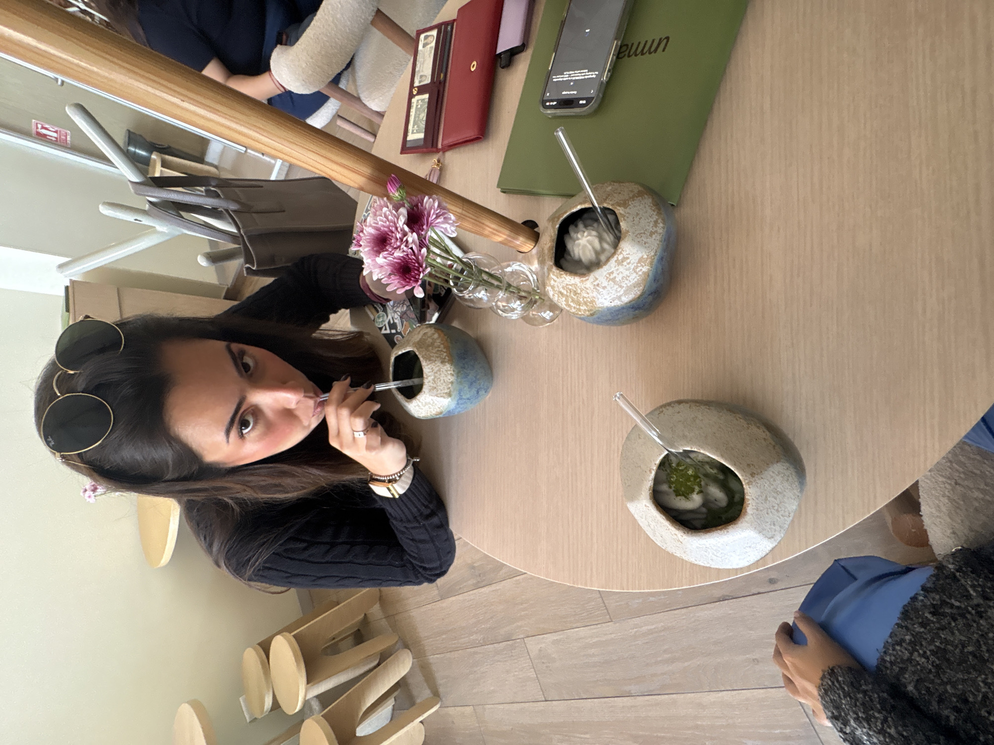 The image depicts an indoor scene within a modern cafe or restaurant in Puebla, Mexico. A woman with dark hair, wearing sunglasses on her head and a black knitted top, is positioned centrally, leaning forward over a light-colored wooden table. She is looking directly at the camera, actively drinking from a clear straw in a blue and beige ceramic cup shaped like a coconut, which contains a green liquid. Her left hand, adorned with a ring and bracelets, holds the straw.
On the same light wooden table, three ceramic coconut-shaped cups are present. One is being used by the central woman. Another, blue and beige, holds a white, frothy substance with a clear straw. The third, light beige, contains a green liquid with white solid elements and a clear straw. A small glass vase containing three pink-purple chrysanthemum-like flowers is centrally placed.
Personal items on the table include a red leather wallet displaying US dollar bills, a smartphone (iPhone) with its screen illuminated, resting on a green cover or menu, and a smaller black item beneath the wallet. A portion of another person's arm, wearing a grey sweater, and blue pants are visible in the bottom left foreground, suggesting another individual is seated at the table. In the background, a third person is partially visible, seated on a white textured chair.
The environment features light wooden plank flooring and cream-colored walls. In the background, minimalist light-colored wooden stools are visible, along with a coat rack supporting a brown tote bag. A doorway in the distant background displays a "No Smoking" sign and a fire extinguisher symbol. A tall, light-colored wooden pole stands vertically near the table's edge. The scene is brightly lit.