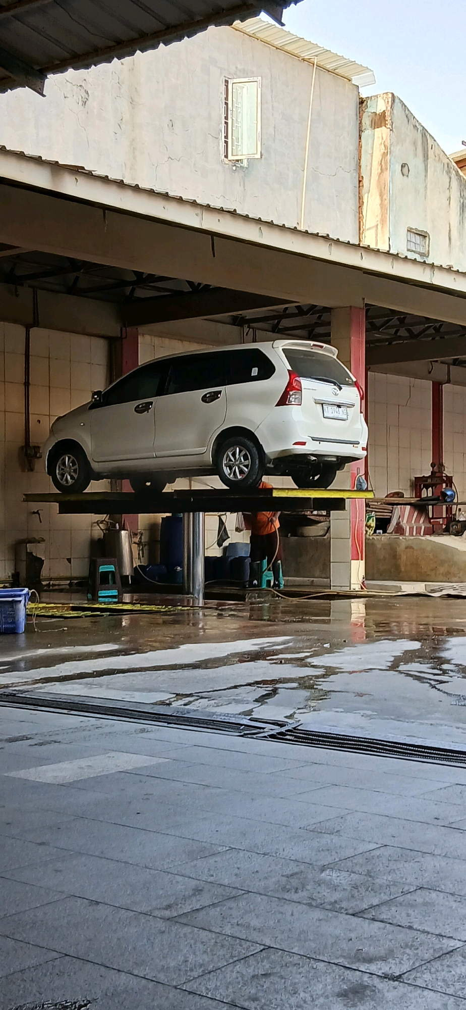 A white minivan is elevated on a car lift at a car wash in Samarinda, Indonesia. The car is positioned at a slight angle, with its right side closer to the viewer. The car wash appears to be an open-air structure with a corrugated metal roof and concrete walls. The ground is wet and reflective, indicating recent activity. In the background, a person in an orange shirt can be seen working under the car, holding a hose. Various cleaning supplies and equipment, including buckets and a large blue barrel, are visible in the vicinity. The overall impression is a functional car wash environment.
