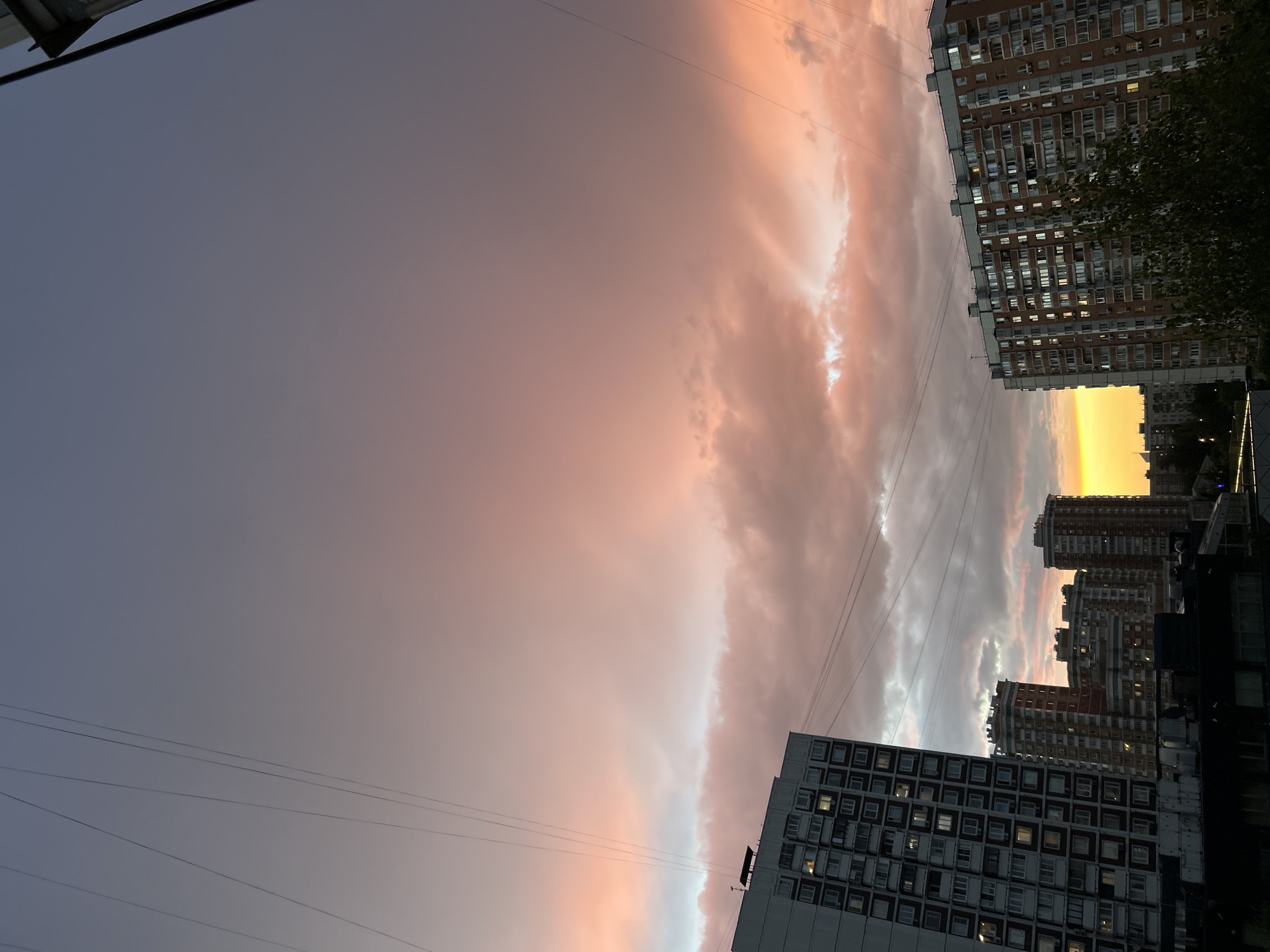 The image depicts a dramatic sunset over a dense urban landscape in Moscow, Russia. The sky is a vibrant canvas of pink, orange, and purple hues, with large, textured clouds dominating the upper portion of the frame. Several tall residential buildings, characteristic of Soviet-era architecture, are visible. The building on the left is a large, grey, multi-story apartment complex with numerous windows. To its right, a cluster of brick-faced apartment buildings rises, some with orange-red facades. In the distance, more buildings and the silhouette of trees can be discerned. Power lines are strung across the sky, adding a geometric element to the scene. The light from the setting sun illuminates the windows of the buildings, giving them a warm glow, and casts a bright yellow light on the horizon. The overall impression is one of urban life bathed in the transient beauty of a colorful sunset.