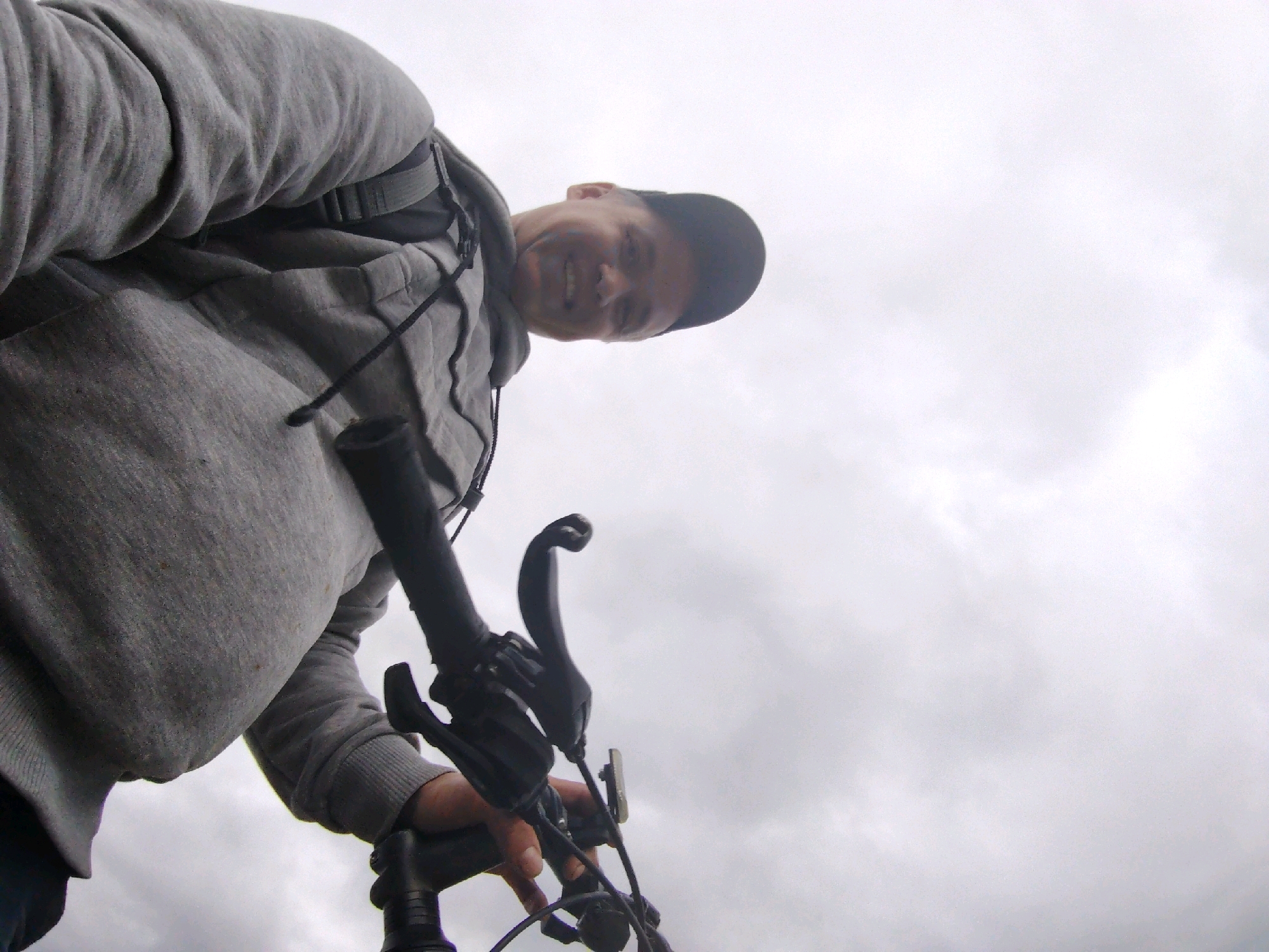 A low-angle, slightly tilted medium shot captures a person from the chest up, holding bicycle handlebars. The person is wearing a gray hooded sweatshirt and a dark baseball cap. Their face, partially obscured by the cap and the angle, is turned towards the camera, with a smile visible. The background is a bright, overcast sky with visible clouds. The bicycle handlebars are black and appear to be made of plastic and metal. The scene is set outdoors, with no specific city landmarks or context visible in the image, therefore, it cannot be definitively placed in Chaikovsky, Russia.