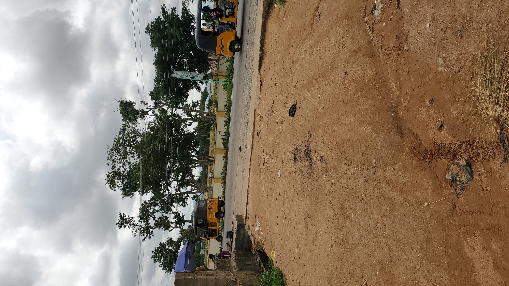 The image depicts a street scene in Jalingo, Nigeria, under a cloudy sky. In the foreground, a dusty, unpaved area with some scattered debris and dry grass. A paved road runs horizontally across the mid-ground, with two yellow auto-rickshaws (locally known as "Keke Napep") visible. The auto-rickshaw on the left is stationary, and two individuals are seated near a blue awning to its left, possibly at a roadside stall. The auto-rickshaw on the right is in motion, with two occupants visible. Beyond the road, a line of trees with dense green foliage borders a low yellow wall, behind which a few buildings and more trees are visible. A signpost with some writing is also present. The overall impression is of a typical urban street in a warm climate.