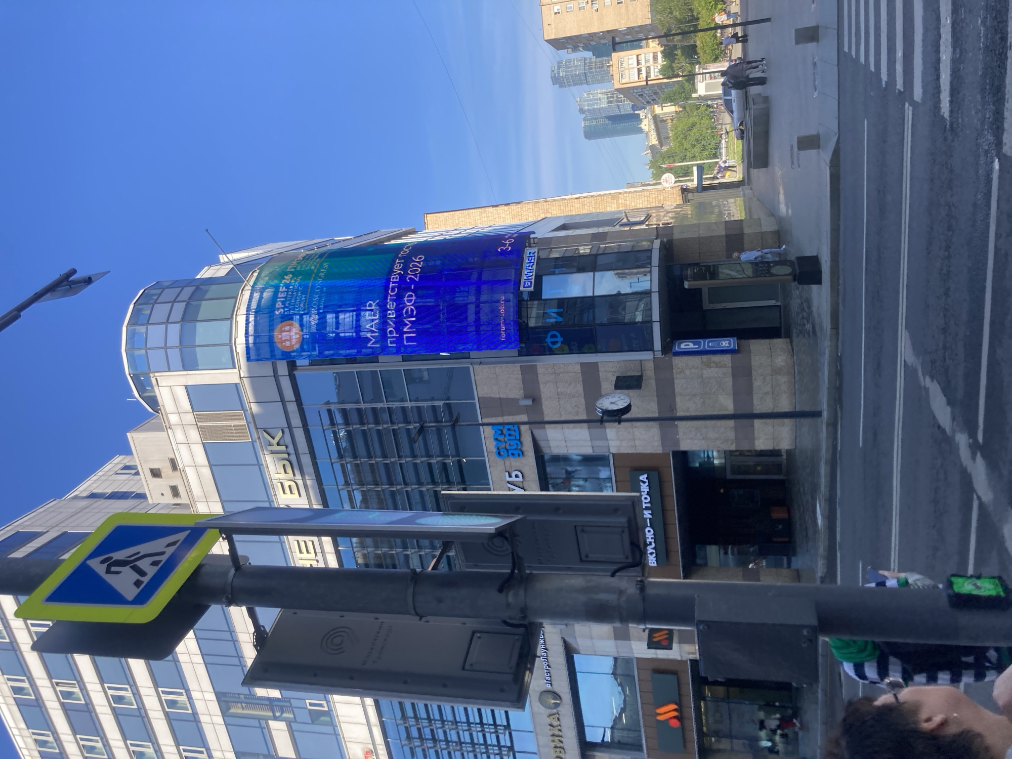 The image displays a bright, sunny daytime scene of a bustling urban street in Moscow, Russia, featuring modern architecture and commercial activity. The central subject is a multi-story building with a distinctive curved glass and steel facade, adorned with a large, vibrant blue LED screen.
On the street level, a pedestrian zebra crossing is visible, indicating an active thoroughfare. A tall street light pole stands in the foreground, featuring a yellow and blue pedestrian crossing sign. Several pedestrians are scattered on the sidewalk and near the street, some distant and blurred, suggesting movement. Other street furniture, including potential kiosks or display panels, are also present.
The prominent LED screen on the building displays text in both Russian and English. Key messages include "MAER приветствует гостей ПМЭФ - 2026" (MAER welcomes guests to SPIEF - 2026), referring to the St. Petersburg International Economic Forum. Additional text related to the event, such as "SPIEF'26 ПМЭФ", "ST PETERSBURG INTERNATIONAL ECONOMIC FORUM", "HOROSCONGRESS", and "forum-spb.ru", along with dates "3-6", are also visible.
Below the LED screen, the building houses several businesses. Identifiable commercial signs include "ВКУСНО-И ТОЧКА" (Vkusno — i Tochka), which is the Russian successor to McDonald's, and "GYM." Other partial signs like "БЫК" (Bull) and "ВИКАТ" are also visible. A classic round clock is mounted on the building's facade. The sky is clear and bright blue, suggesting fair weather.