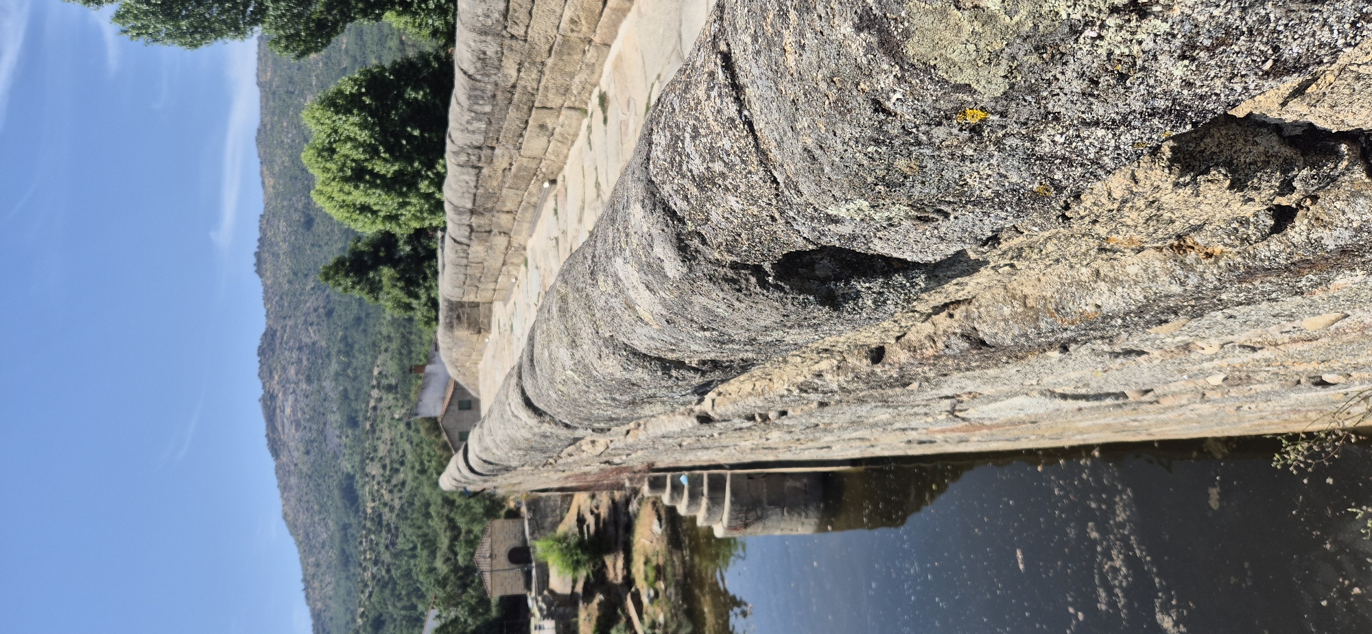 The media file is a photograph taken outdoors, featuring a stone aqueduct or wall alongside a body of water. The scene is dominated by the weathered texture of the stone, which appears aged and possibly covered in lichen, with visible layers and cracks. The perspective is low and angled, looking across the top of the stone structure towards a landscape of green, tree-covered hills under a clear blue sky with a few wispy clouds.
In the background, glimpses of buildings or structures can be seen nestled within the foliage on the hillside. The water in the foreground reflects the sky and the aqueduct, with some debris or plant matter floating on its surface. The lighting suggests it is daytime, likely sunny, given the bright sky and clear visibility of the landscape.
The focus is primarily on the natural and man-made elements of the setting: the rough, organic texture of the rock and the engineered structure of the aqueduct, juxtaposed with the wildness of the surrounding hills and sky. There is no discernible activity or event taking place, and the image conveys a sense of stillness and natural beauty. No text is visible in the image.