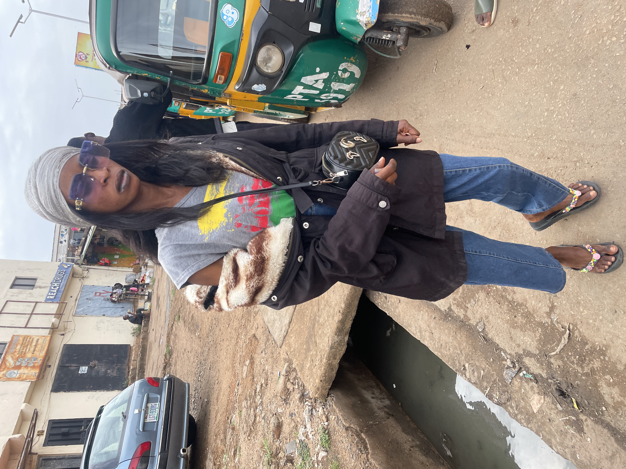 A woman stands on a street in Jos, Nigeria, posing for a photo. She is wearing a grey head covering, sunglasses, a grey T-shirt with red, yellow, and green colors, a black jacket with a white faux fur lining, blue jeans, and sandals. A small black purse hangs from her shoulder. In the background, a green and yellow auto rickshaw is partially visible. Further back, a parked blue car with a license plate number LG 930 PG can be seen in front of a building that has a sign that says "REACH DRUGS" on it. The ground appears to be a concrete sidewalk or street that is cracked and worn. A drainage ditch runs alongside the street. There is evidence of litter and dirt on the ground. In the distance, people are sitting or standing outside a building. The sky is overcast.