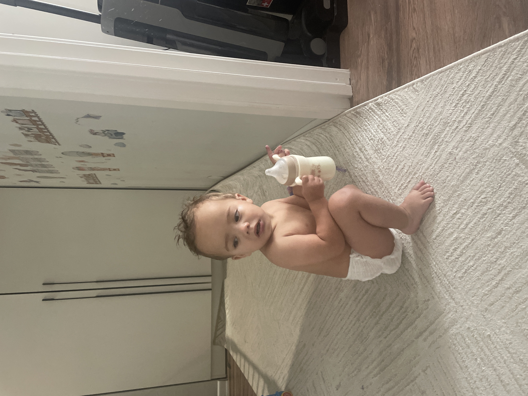A toddler wearing a diaper sits on a rug in what appears to be a home. The toddler is holding a baby bottle. Behind the toddler, there are closed cabinet doors and a wall with decals. To the right, a doorway leads to another room, where a vacuum cleaner is visible. The flooring consists of a rug and wood laminate. The scene is indoors, and there is no indication of the city of Almaty, Kazakhstan.