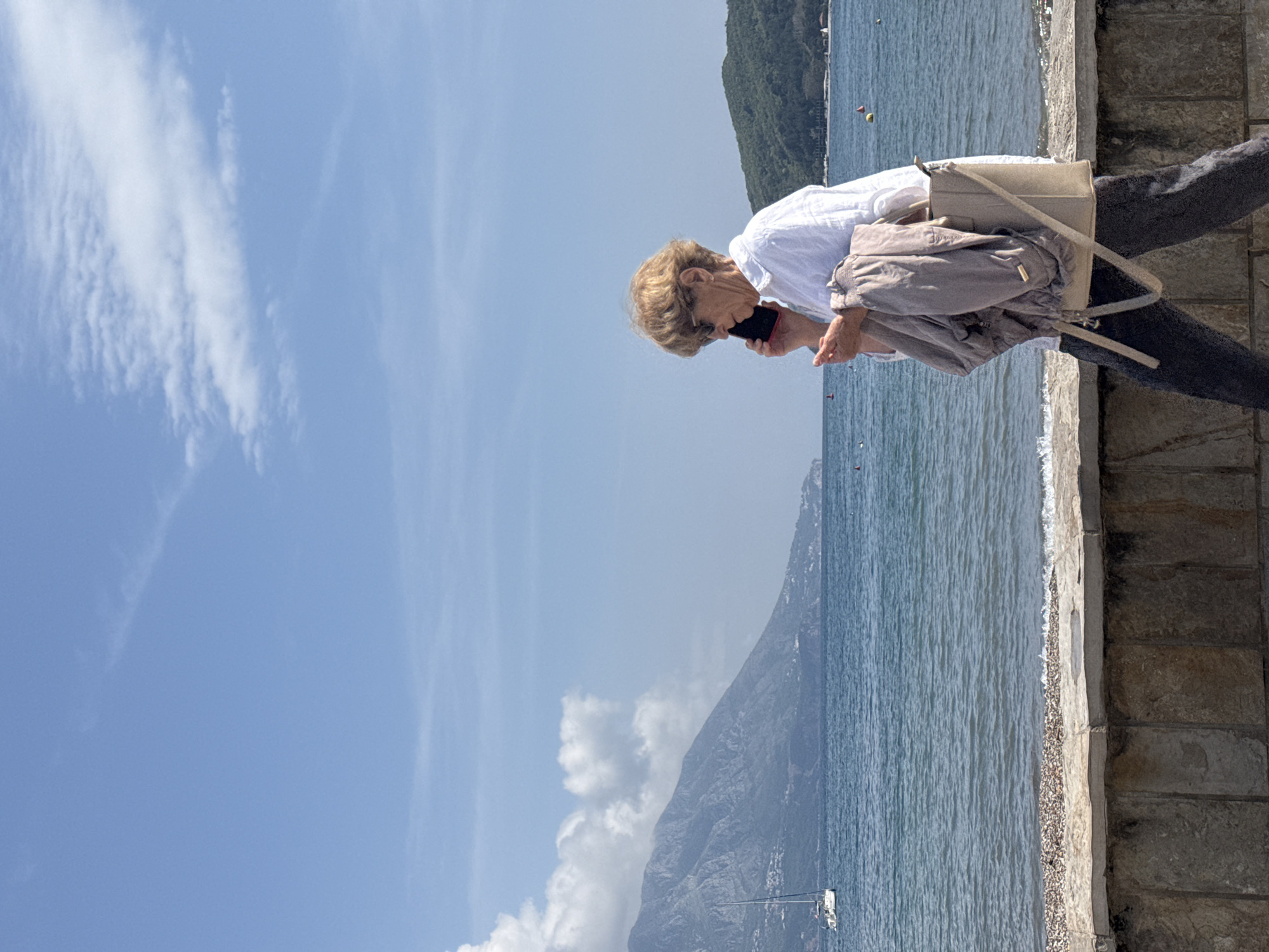 The image captures a bright, sunny scene in Budva, Montenegro, depicting a woman standing on a stone promenade beside the sea. The perspective is unusual, with the image rotated 90 degrees clockwise, making the sky appear on the left and the water at the bottom, relative to the main subject.
The primary subject is a woman, likely middle-aged or older, with short, light brown hair, positioned on the right side of the frame. She is wearing a white long-sleeved shirt and dark trousers. A beige shoulder bag is slung over her shoulder, and a light-colored jacket is draped over one arm. Her head is bowed, and she holds a smartphone with a red or dark case to her ear, appearing to be engaged in a phone call or intently listening.
The setting is a picturesque seaside location. A sturdy stone wall or promenade runs horizontally (when the image is correctly oriented) across the foreground, separating the viewer from the water. Below the wall, a narrow strip of pebbly shore meets the calm, blue-green water. In the background, across the water, large, rocky mountains rise under a clear, expansive blue sky. Some scattered white clouds are visible, particularly on the left side of the sky. A small white boat and a few colored buoys are sparsely dotted in the water. The overall impression is one of leisure and communication in a scenic coastal environment during daylight hours. No visible text is present in the image.