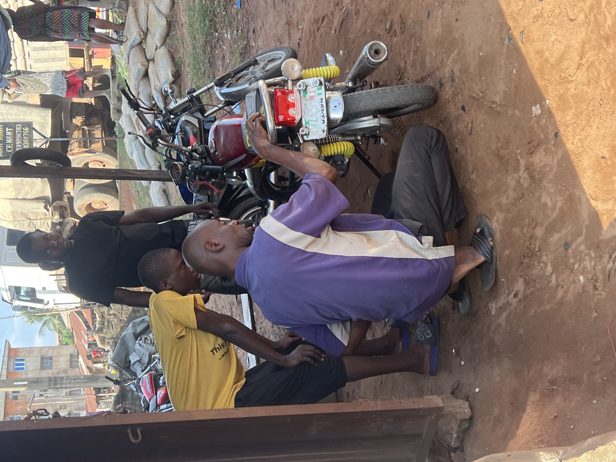 In Nkpor, Nigeria, a group of three men are gathered around motorcycles on a dusty, unpaved ground. The man in the foreground, wearing a purple and white shirt and dark pants, is kneeling and appears to be working on the rear of a red motorcycle, his hand touching the seat. To his left, a man in a yellow t-shirt and black shorts is bending over, looking down at something near the motorcycle, with his hands extended. To the left of this man, a third man in a black t-shirt stands upright, observing the scene. In the background, a large white truck is parked, with bags of cement stacked nearby. The environment suggests a busy roadside or workshop area, with buildings and debris visible.